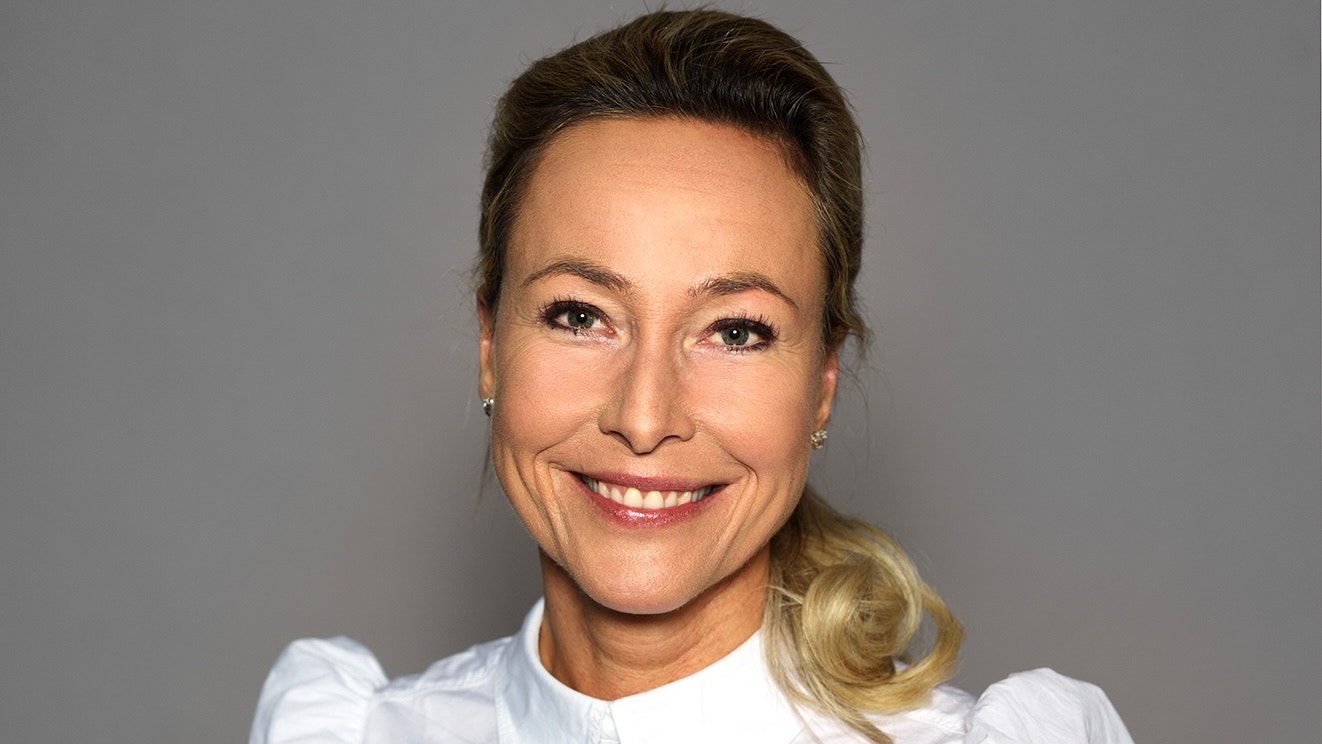 Porträttfoto av en kvinna med blont hår som ler mot kameran. Hon bär en vit blus mot en grå bakgrund.