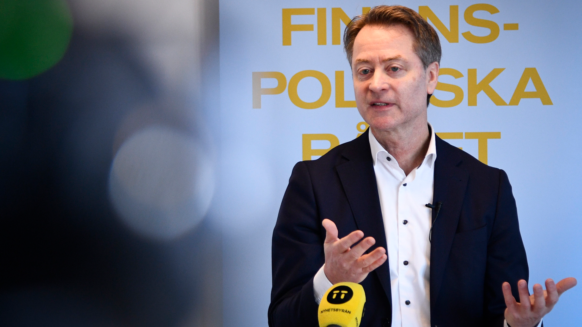 En man i mörk kavaj och vit skjorta talar vid en mikrofon framför en blå bakgrund med gul text.