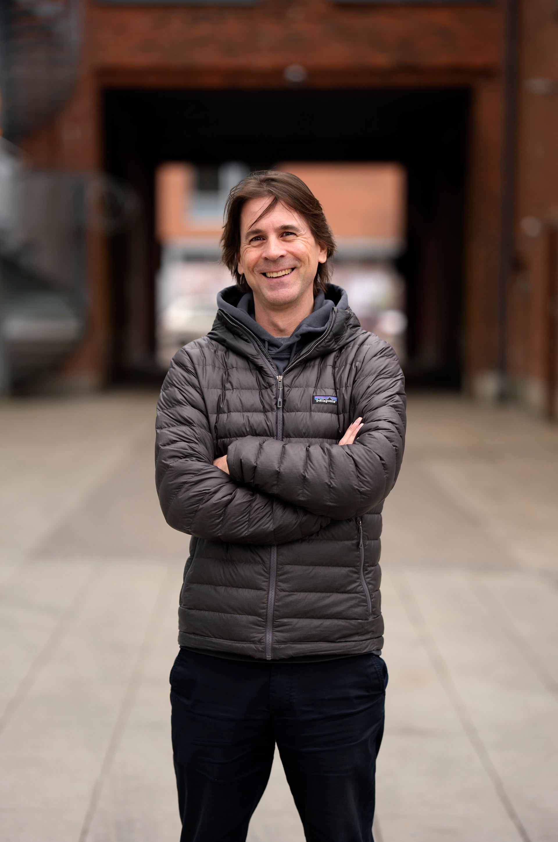 Porträtt av Jan Erik Solem som står utomhus framför en byggnad. Han är klädd i en svart vinterjacka och mörkblå byxor, och ler mot kameran med armarna i kors.