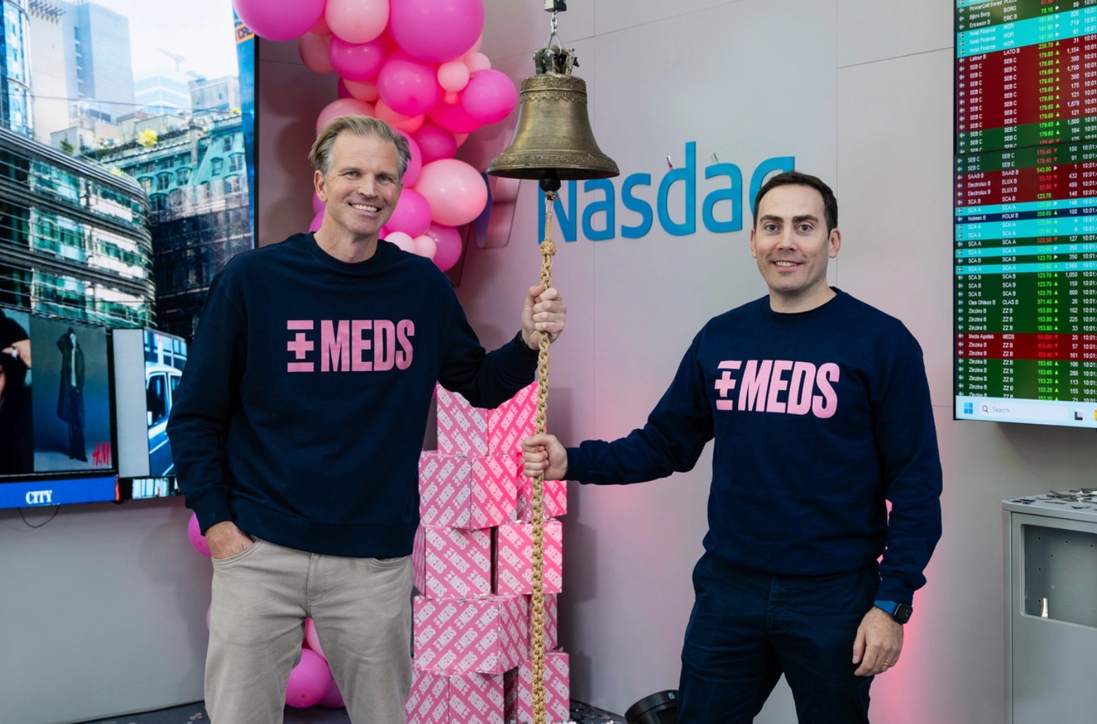 Två personer i svarta MEDS-tröjor håller i en stor klocka vid Nasdaq-börsen. De är omgivna av rosa ballonger och börskurser syns i bakgrunden.