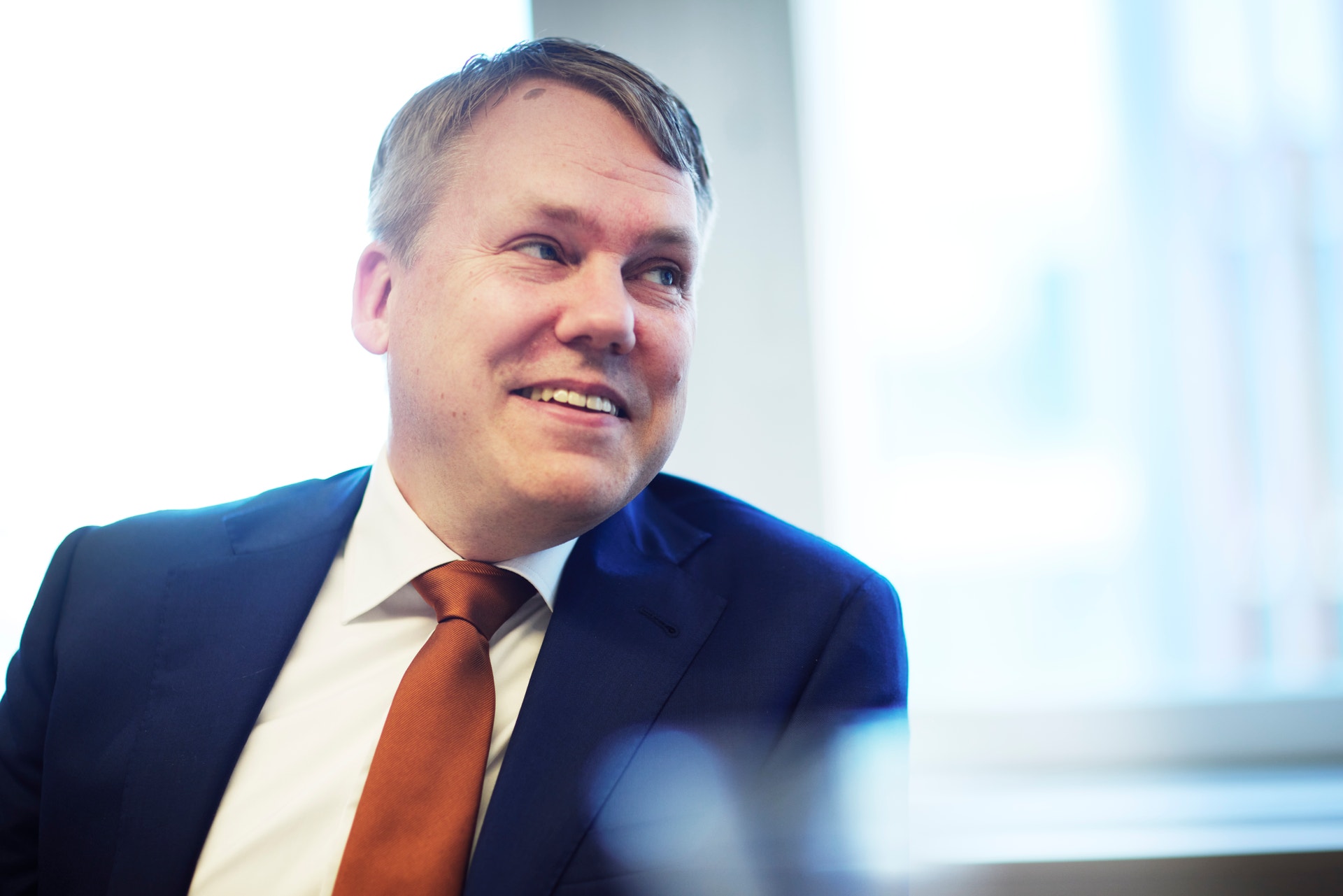 Porträtt av en man i mörkblå kostym och röd slips som ler mot kameran i en ljus kontorsmiljö.