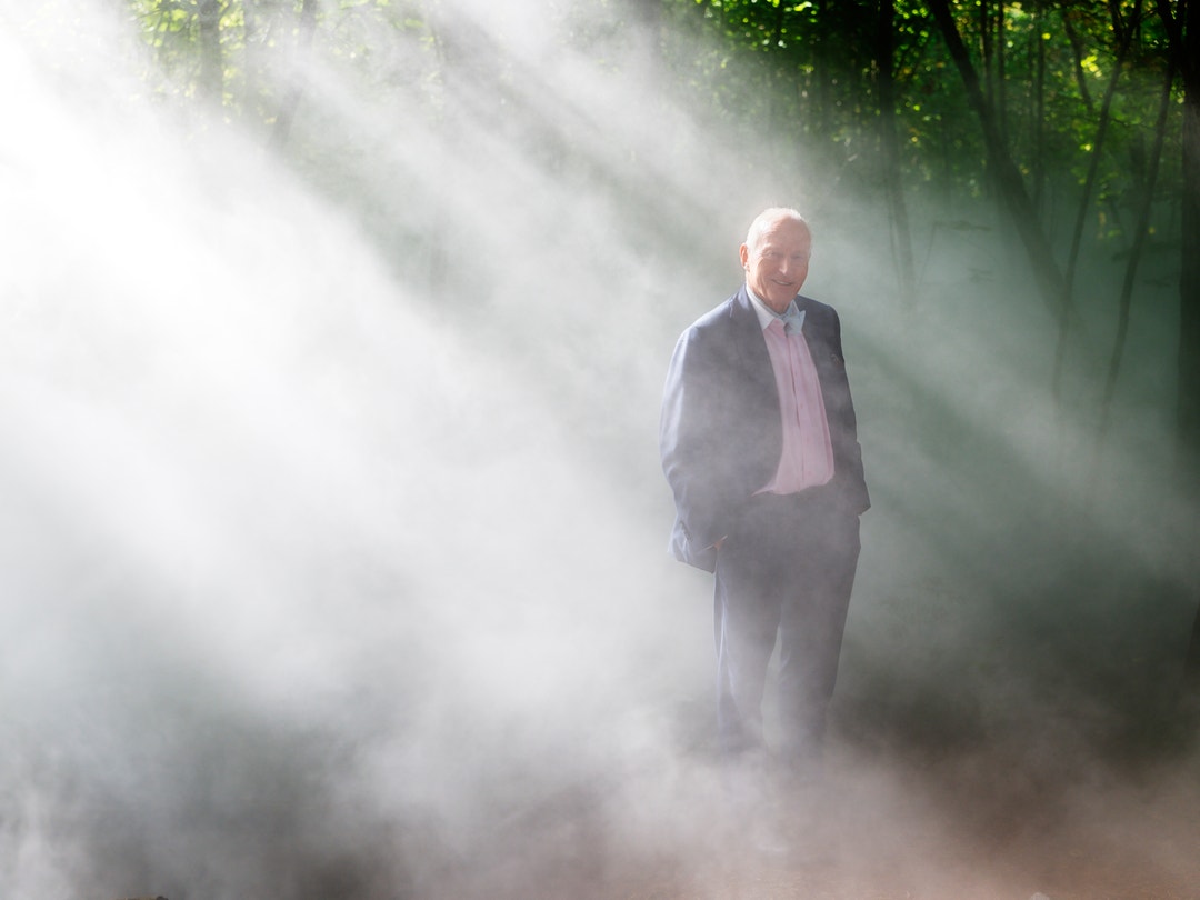 En äldre man i kostym står i en skog omgiven av dimma, med solstrålar som lyser upp luften omkring honom.