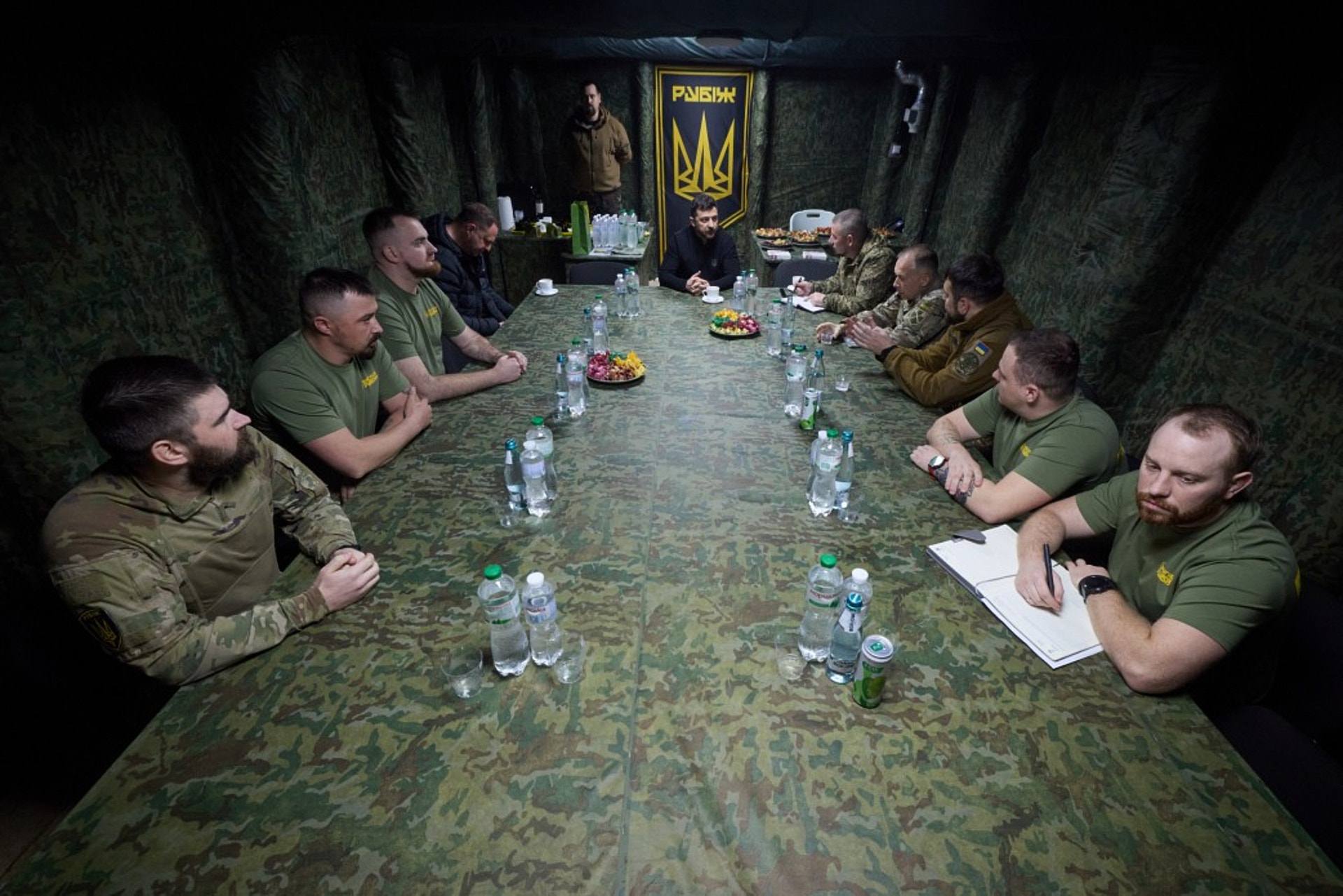 Grupp av soldater i militär uniform sitter runt ett långbord under ett möte i ett underjordiskt rum. Vattenflaskor och mat står på bordet.