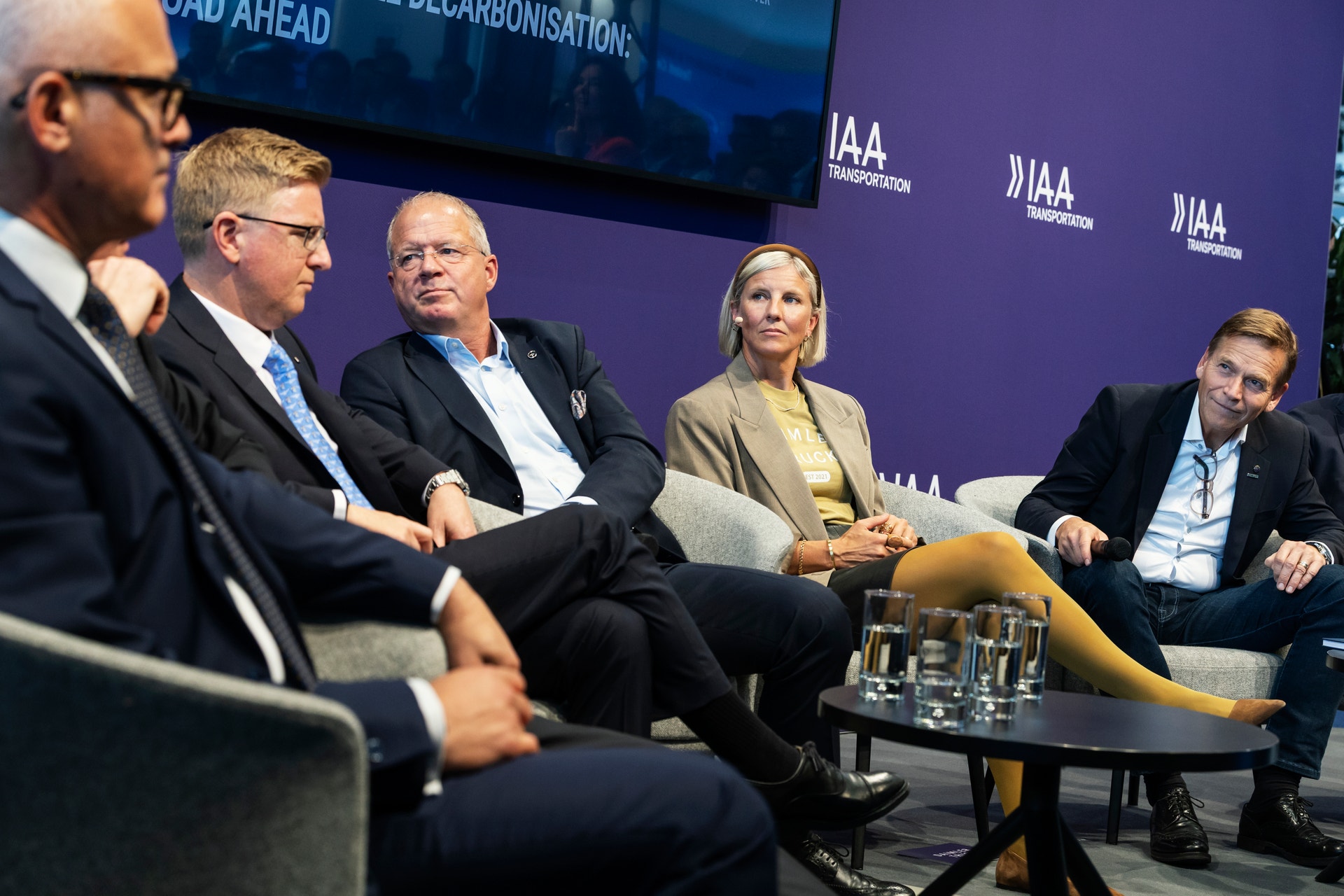 Fyra män och en kvinna sitter i fåtöljer på en scen och samtalar i en paneldiskussion, med IAA Transportation-logotyper i bakgrunden.