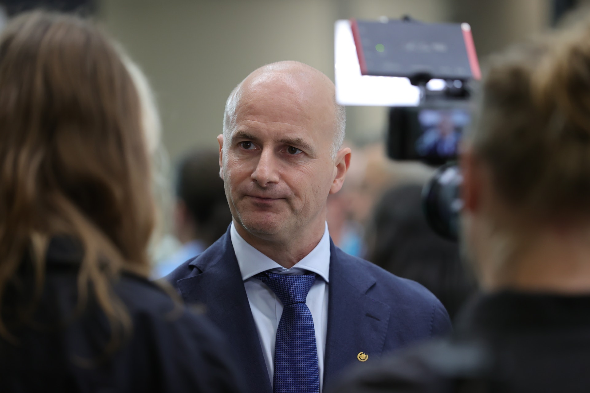 En kalhövdad man i blå kostym och slips blir intervjuad av journalister, varav en håller en filmkamera.