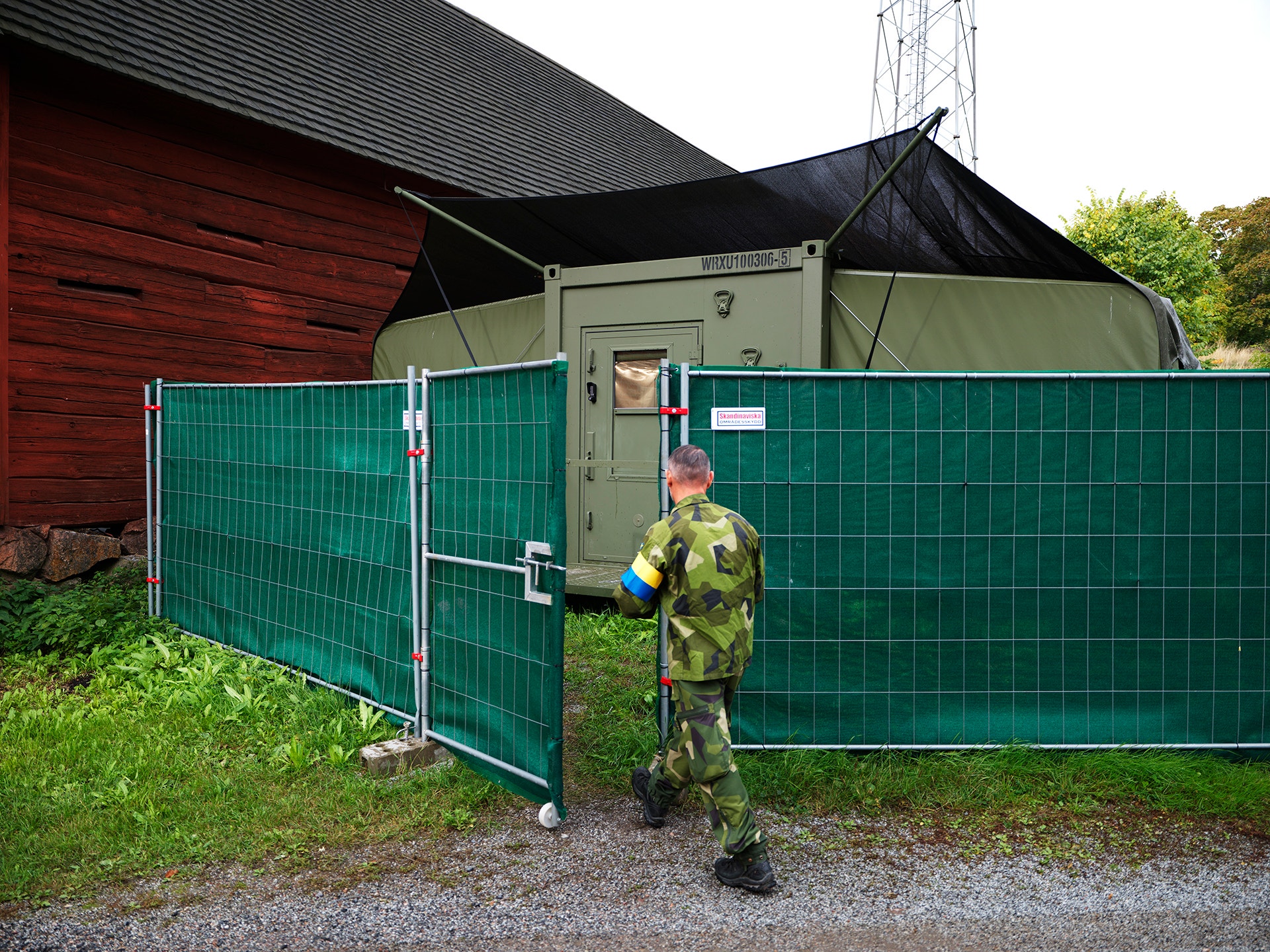 En soldat i kamouflageuniform går genom en grön grindöppning in till ett militärt fältläger med en container och en röd lada i bakgrunden.
