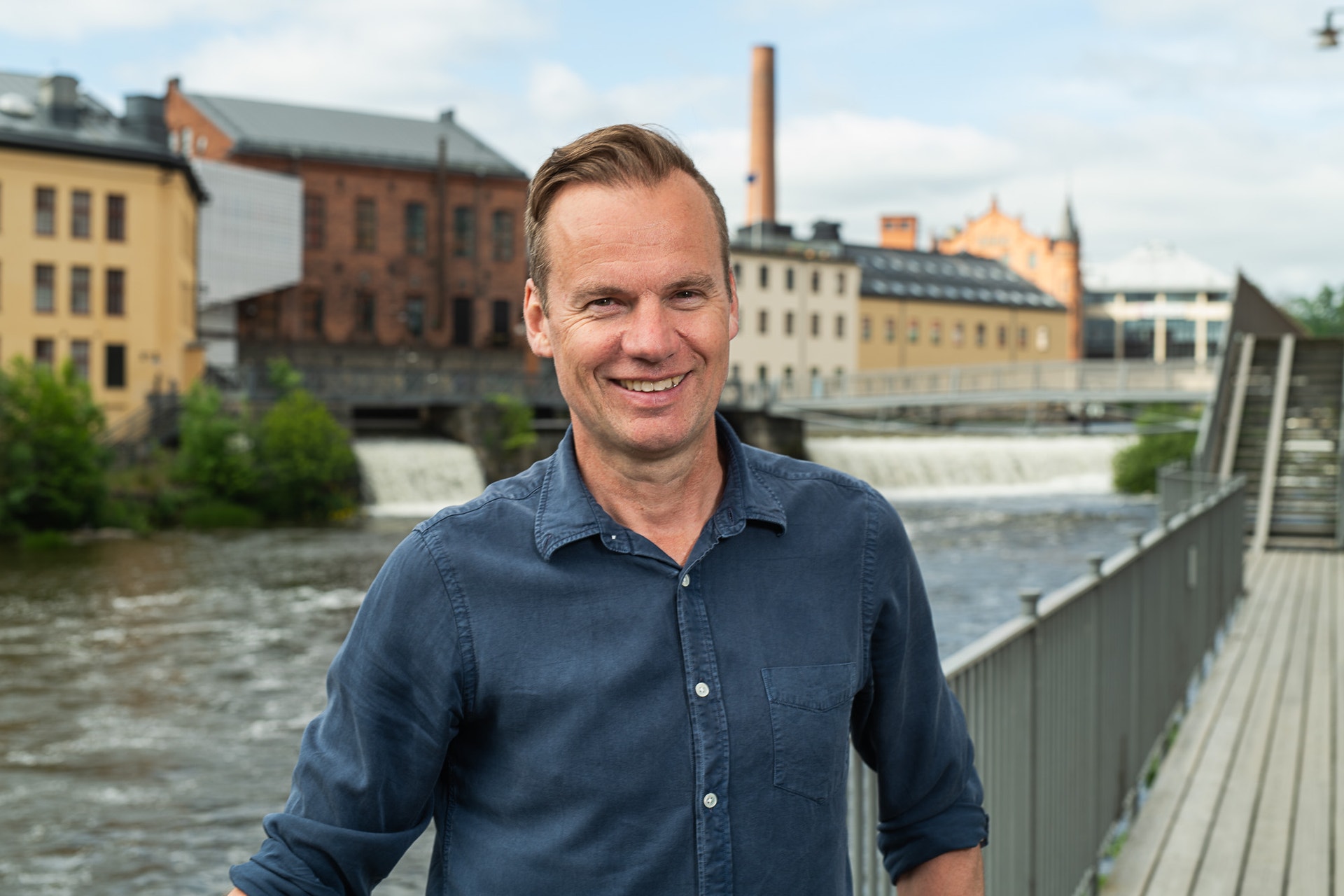 En leende man i blå skjorta står vid ett vattendrag i en stad med äldre industribyggnader i bakgrunden.