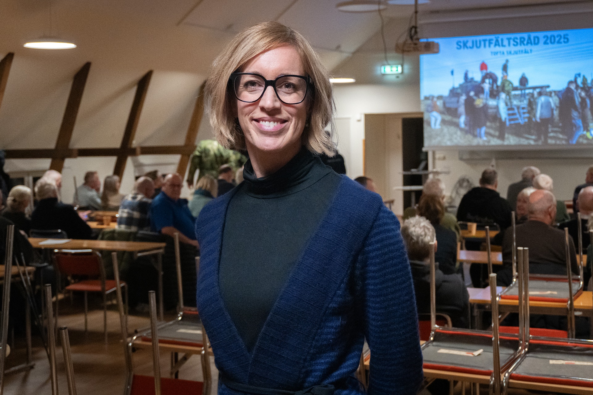 Porträtt av en kvinna med glasögon och blont hår, klädd i mörkblå tröja, som står i ett evenemangsutrymme med andra människor i bakgrunden.