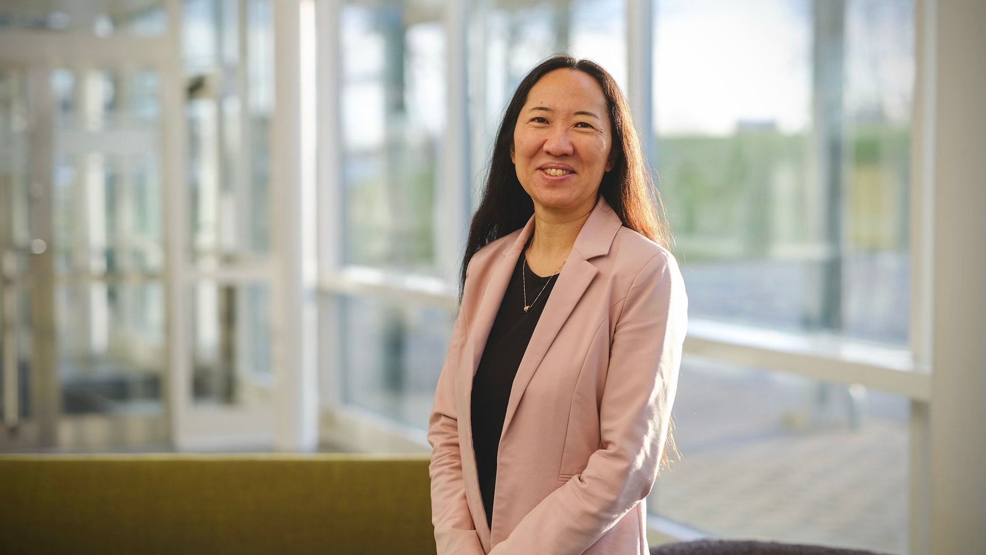 Porträtt av en kvinna i rosa blazer i ett modernt kontor med stora fönster i bakgrunden.