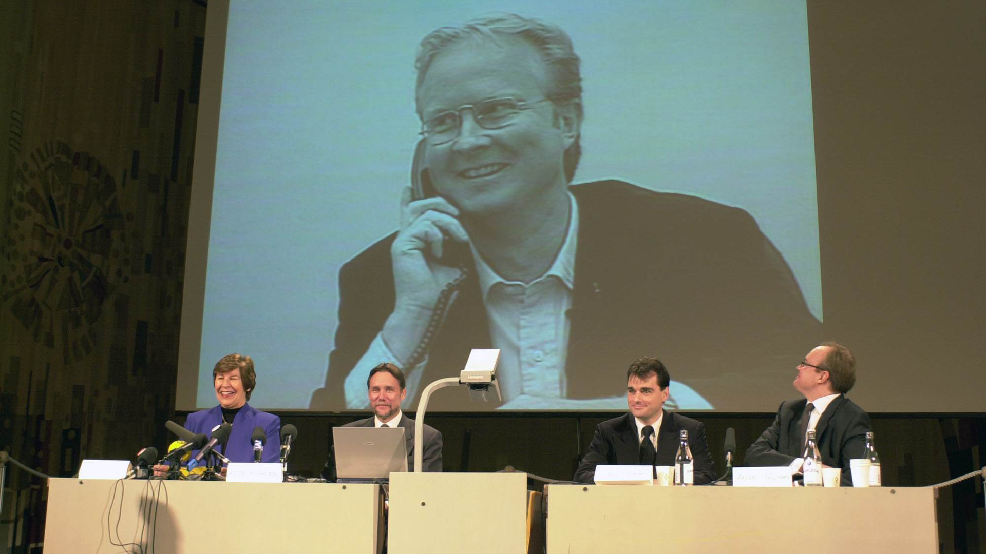 Fyra personer sitter vid ett panelbord under en presentationsskärm som visar en man i glasögon och skjorta.