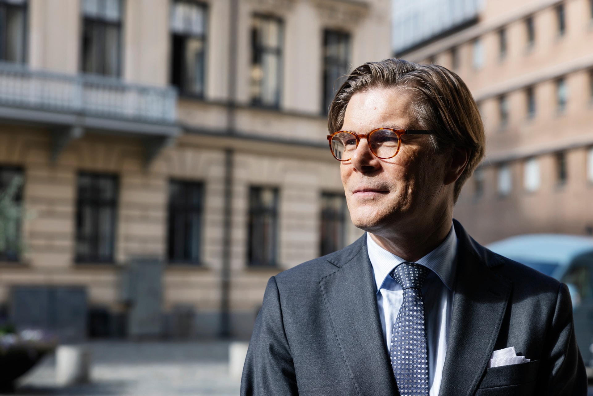 Porträtt av en man i mörk kostym och glasögon som står framför en klassisk byggnad.