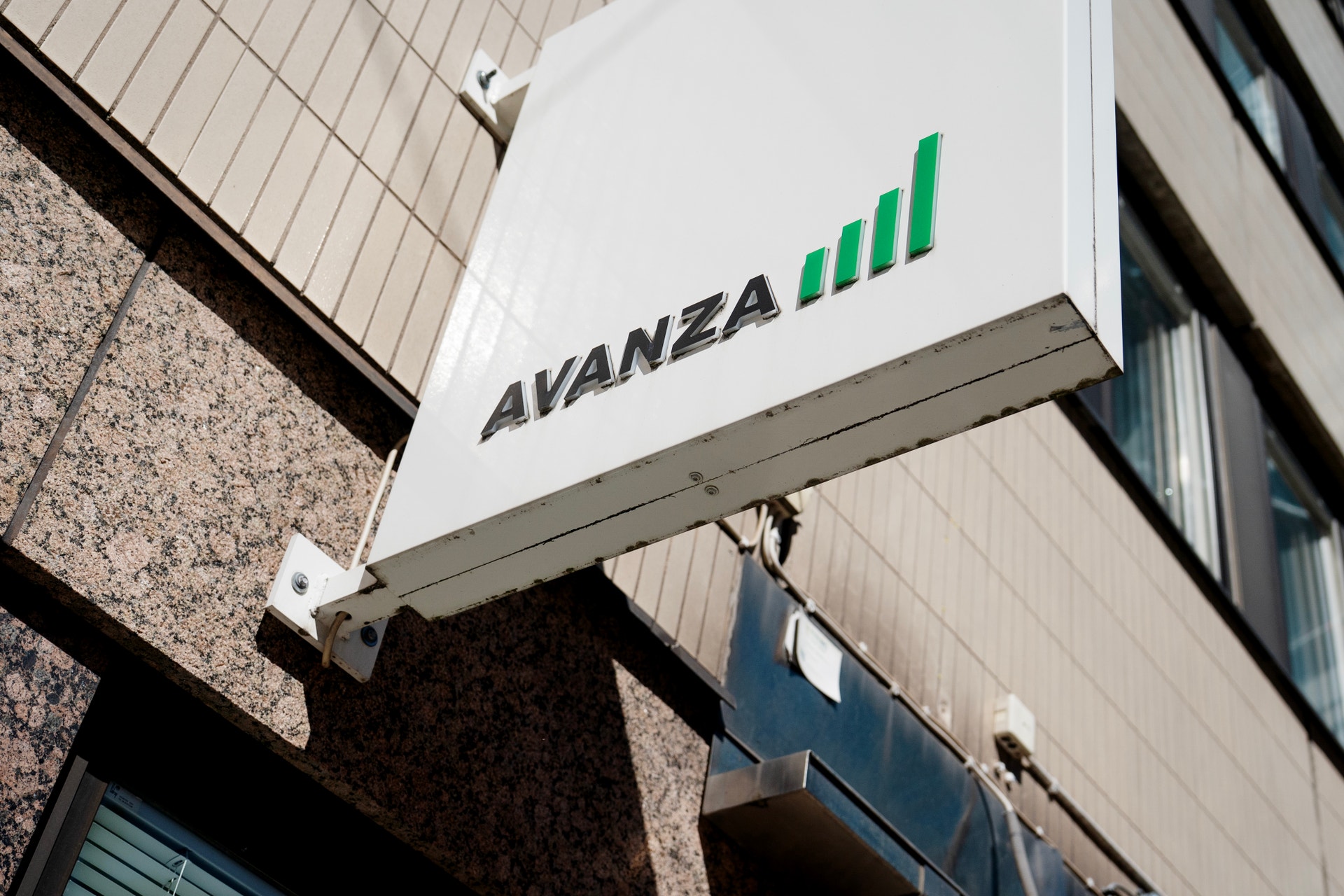 Vit skylt med texten Avanza och en grön stapeldiagram-logotyp monterad på en byggnadsfasad.
