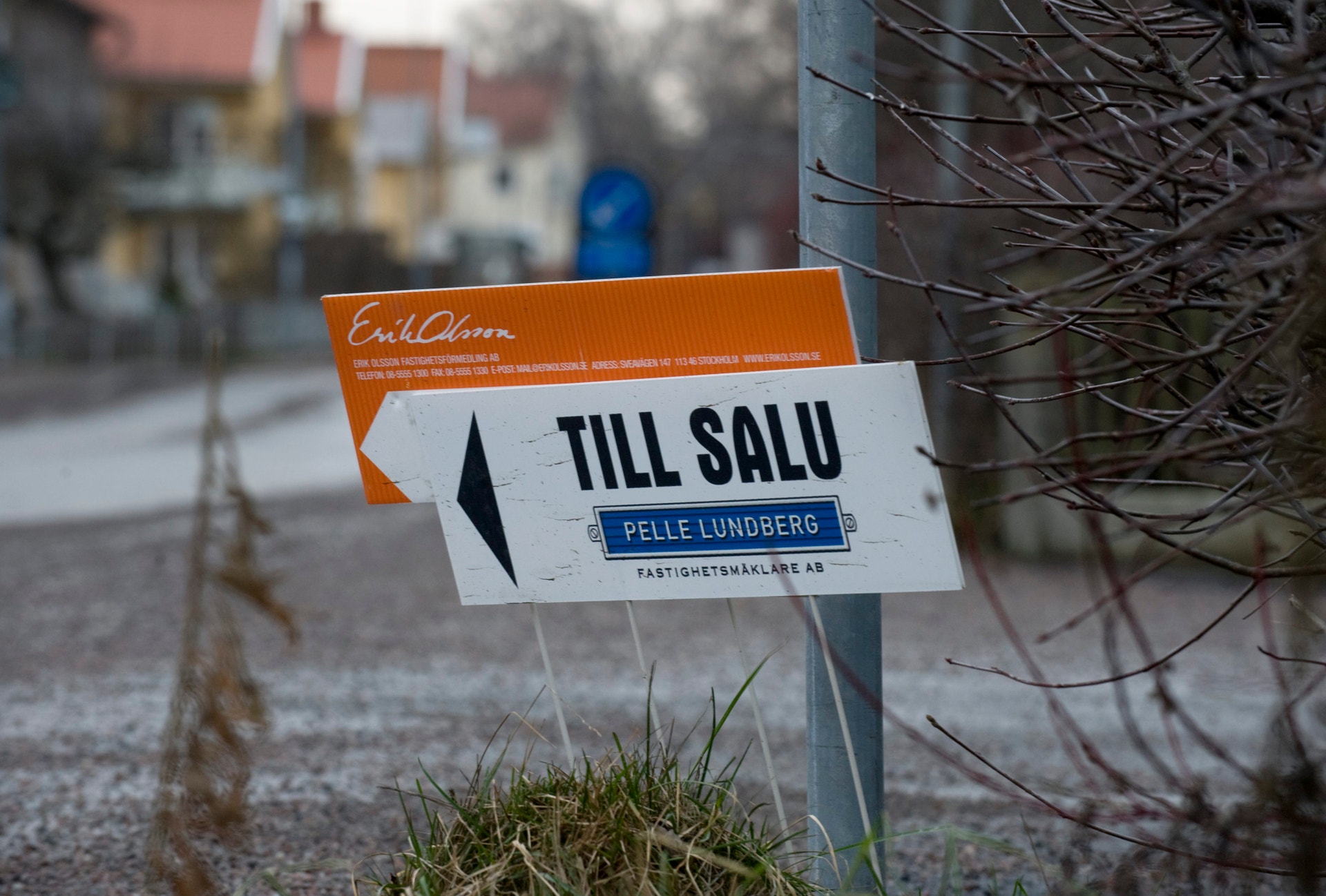 En försäljningsskylt fäst vid en stolpe på en snöig gata med hus i bakgrunden.