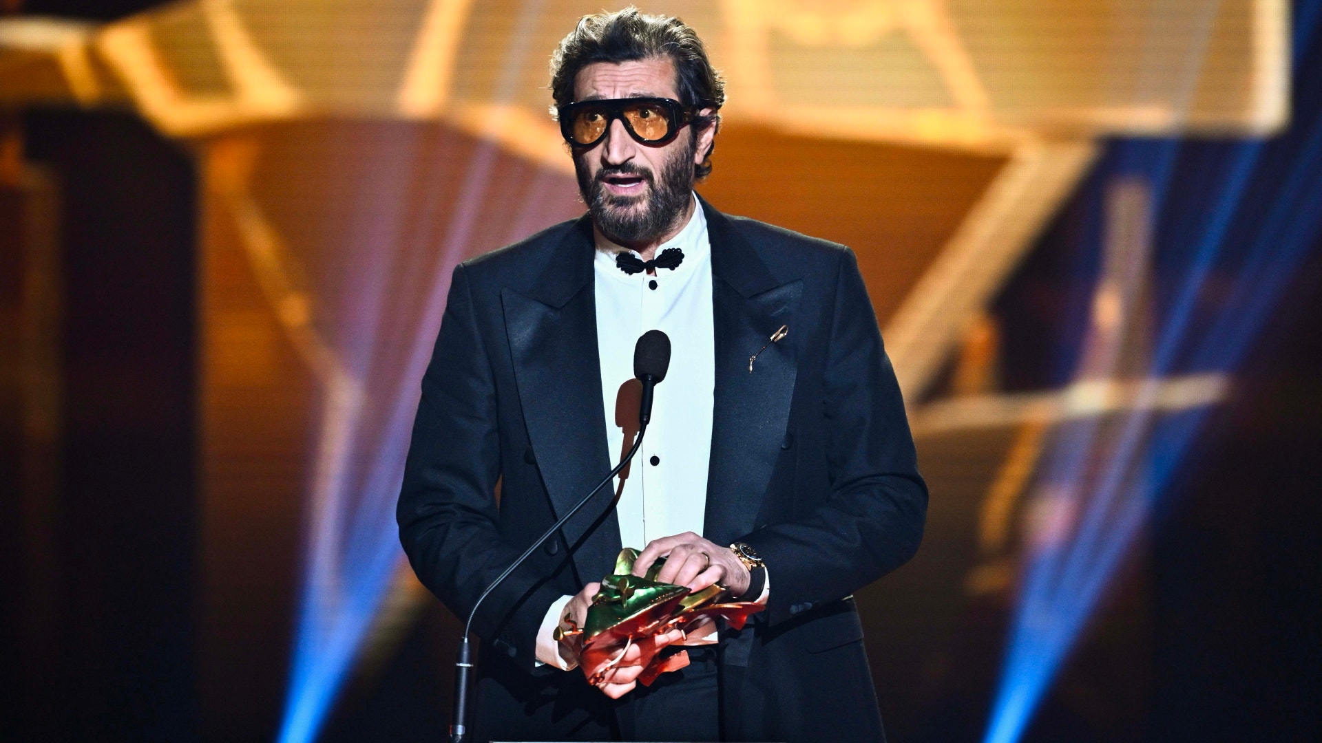 Josef Fares står på en scen vid en mikrofon, klädd i svart smoking med vit skjorta och fluga. Han bär solglasögon och håller i ett rött pris eller utmärkelse. Scenen är belyst med gul och blå belysning i bakgrunden.