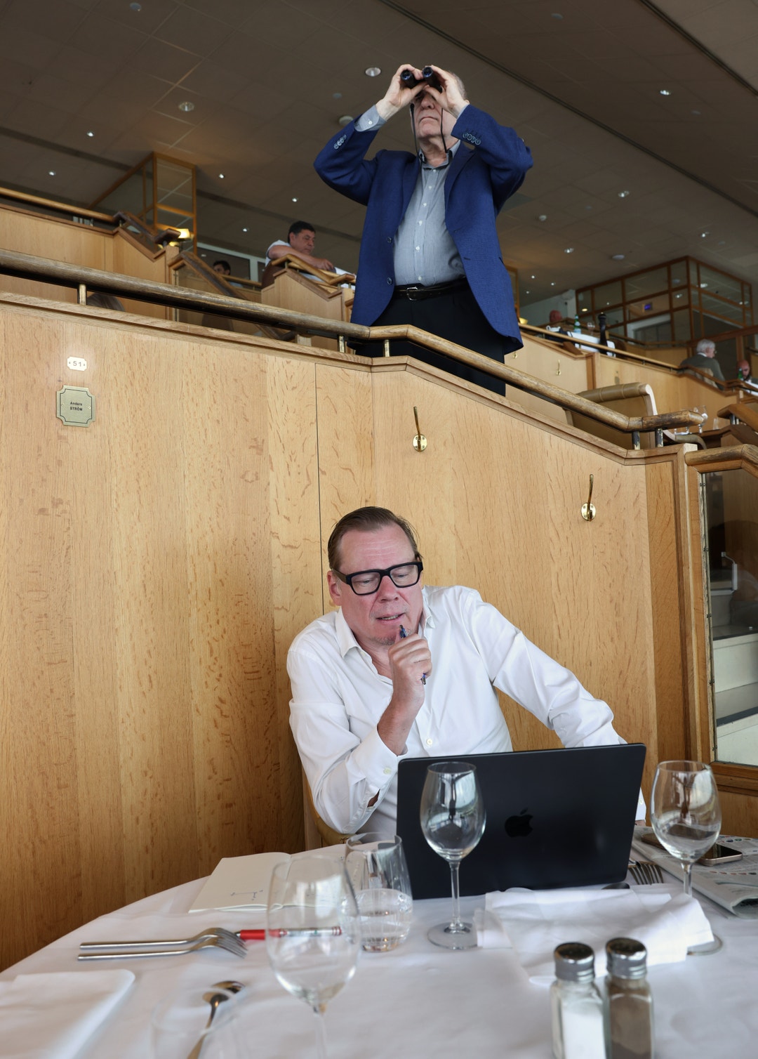 Två personer i en restaurang. En man i vit skjorta sitter vid ett bord med en laptop. En annan person i blå kavaj står på en balkong ovanför och håller händerna på huvudet.