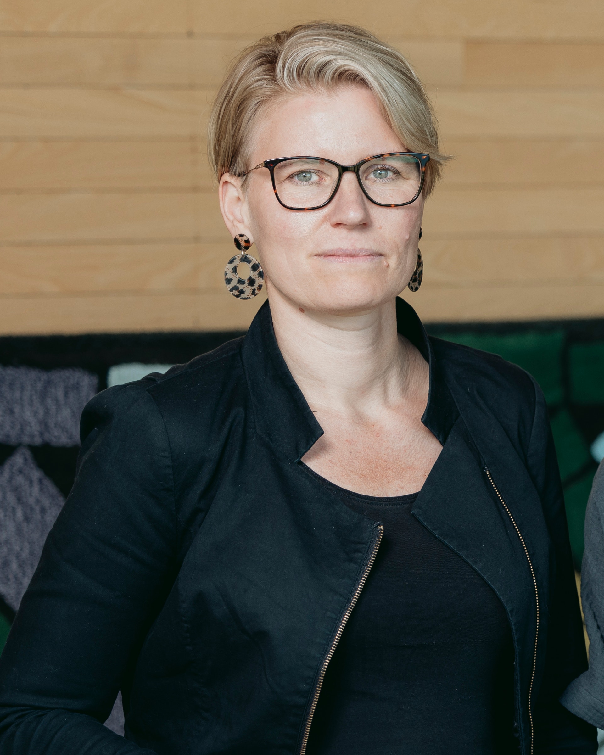 Porträtt av en kvinna med kort ljust hår och glasögon, iklädd svart jacka.