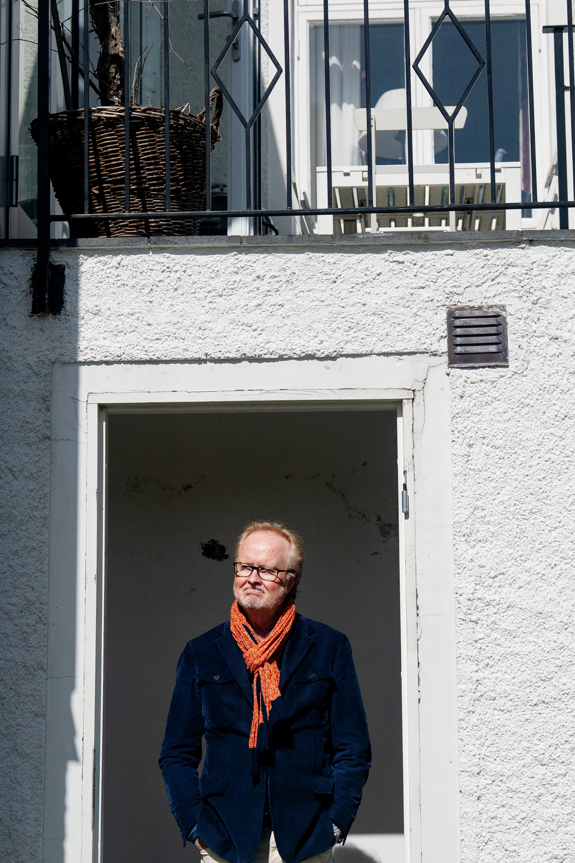 En äldre man med glasögon står i en vit dörröppning. Han bär en mörkblå jacka och ett orangerödt halskläde. Bakom honom syns en modern vit byggnad med stora fönster.