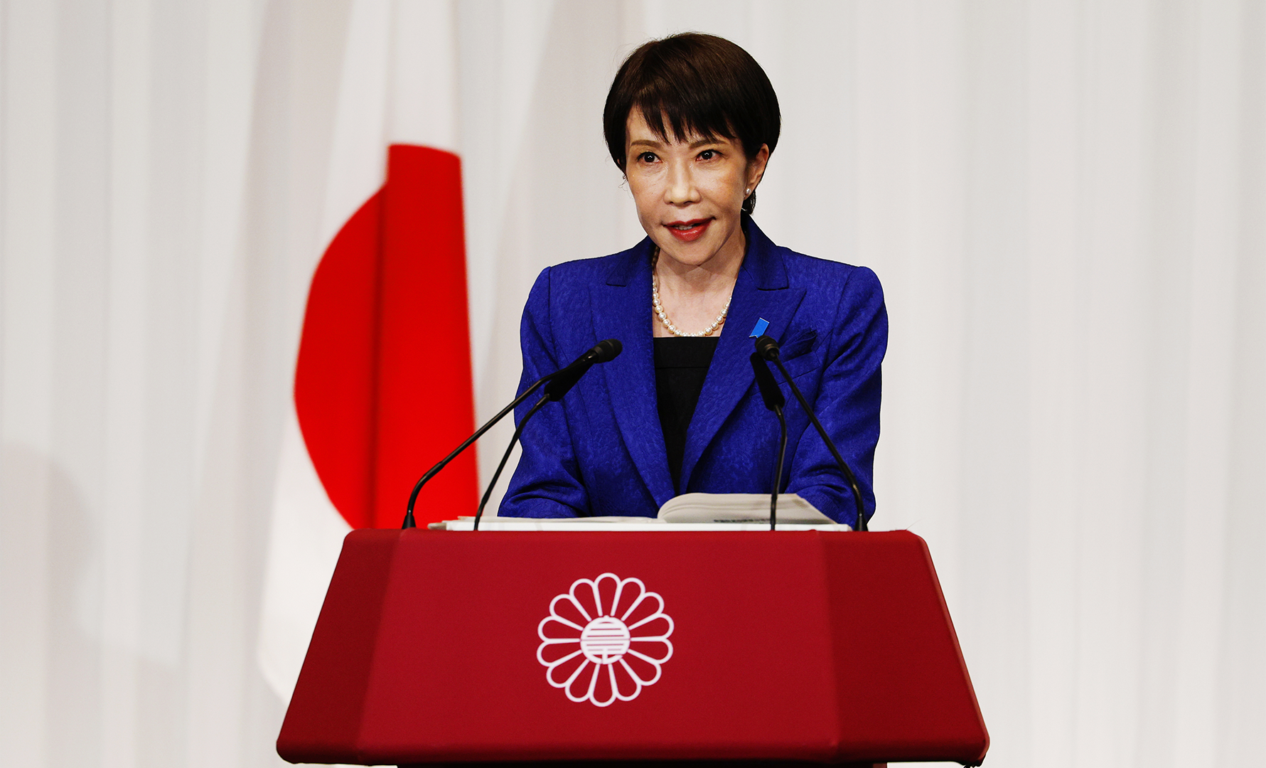 Kvinna i blå kavaj talar vid ett podium med Japans flagga i bakgrunden