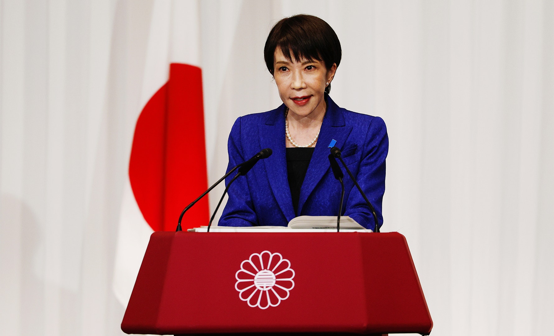 Kvinna i blå kavaj talar vid ett podium med Japans flagga i bakgrunden