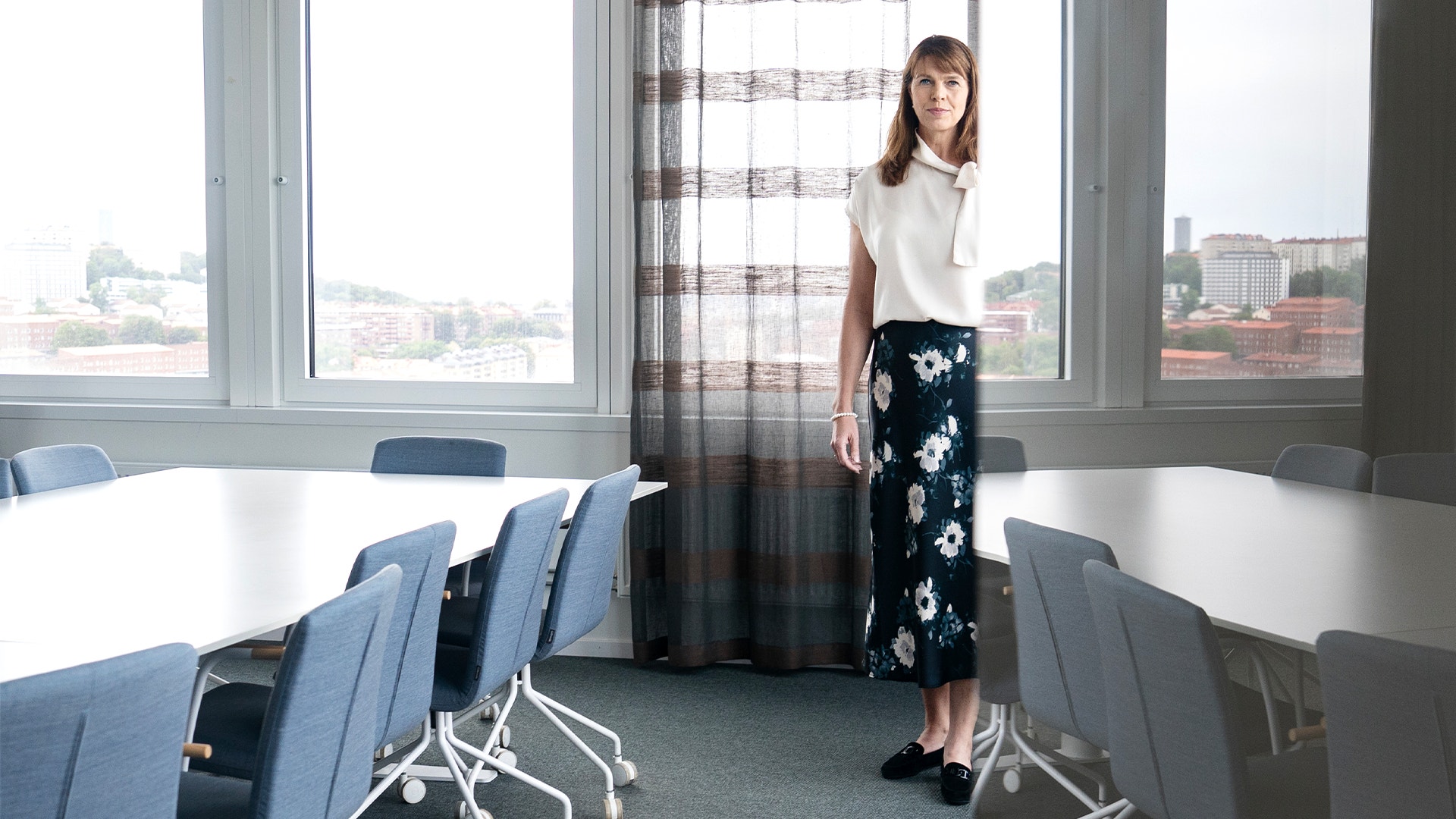 Linda pålsson står i ett av konfferensrummen på sitt kontor. Hon bär blommig långkjol. 