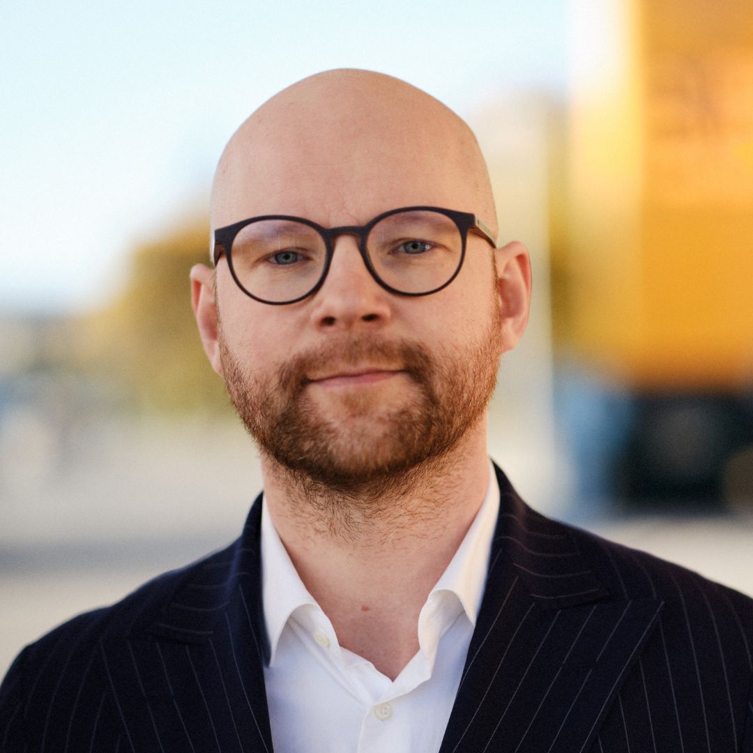 Porträttfoto av en man med glasögon och skägg, klädd i kostym och vit skjorta.