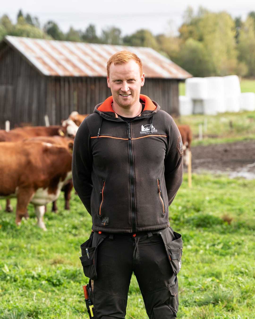 Man med rött hår står på en gård framför bruna kor och lantbruksbyggnader. Han bär en svart och orange hoodie och ler mot kameran.
