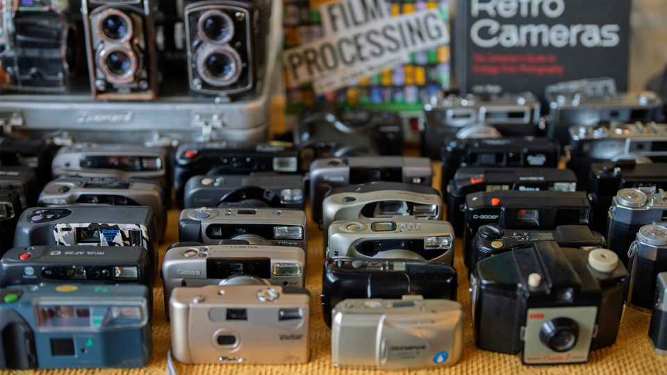 Ett tiotal äldre kameror står uppradade på en hylla i en secondhand-butik.