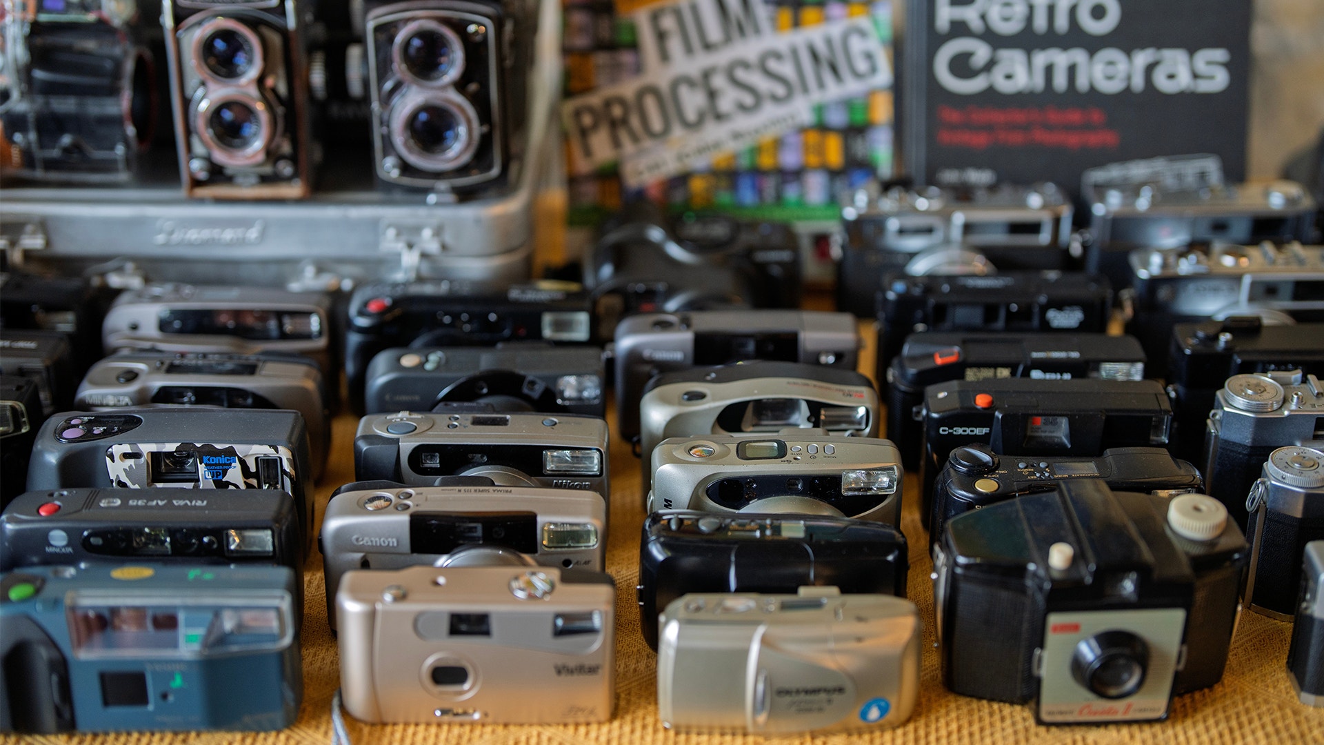 Ett tiotal äldre kameror står uppradade på en hylla i en secondhand-butik. 