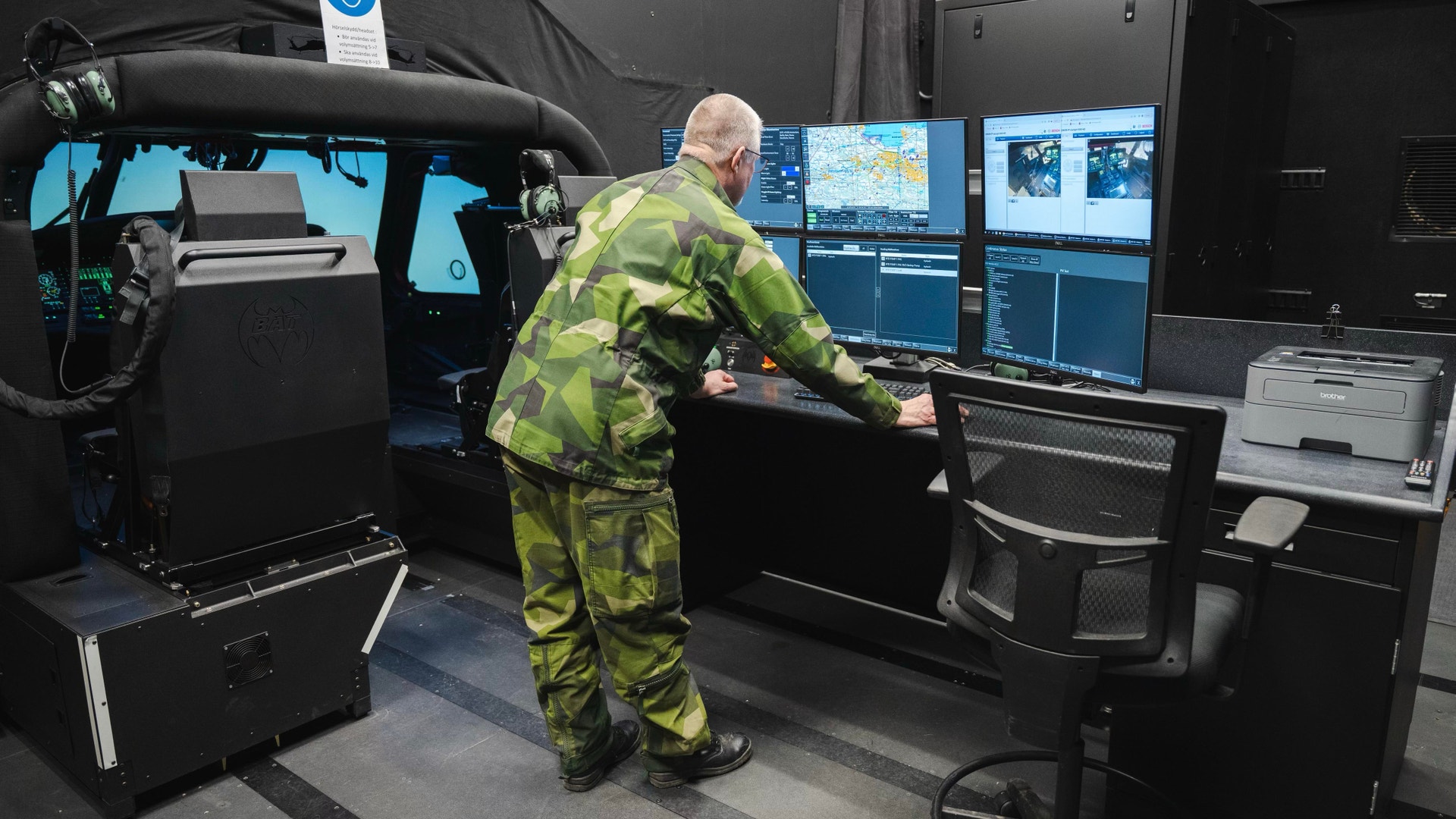 Helikopterpilot i gul-grön flygdräkt arbetar vid en kontrollpanel med flera skärmar som visar kartor och flygdata.