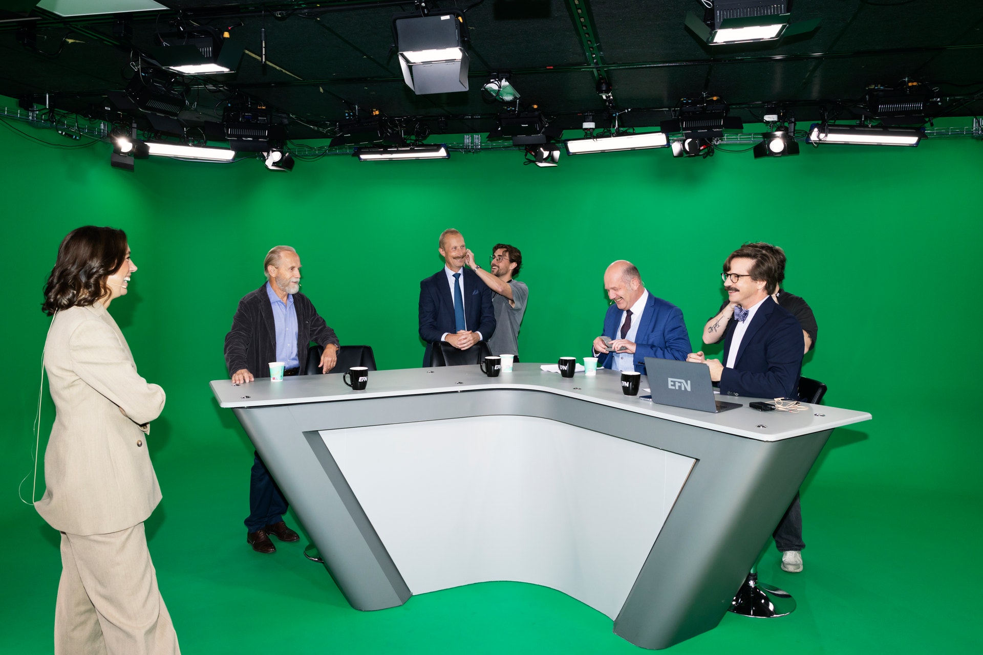 Fyra män i kostym sitter runt ett skrivbord i en tv-studio med grön bakgrund, medan en kvinna står bredvid och en person justerar mikrofon.