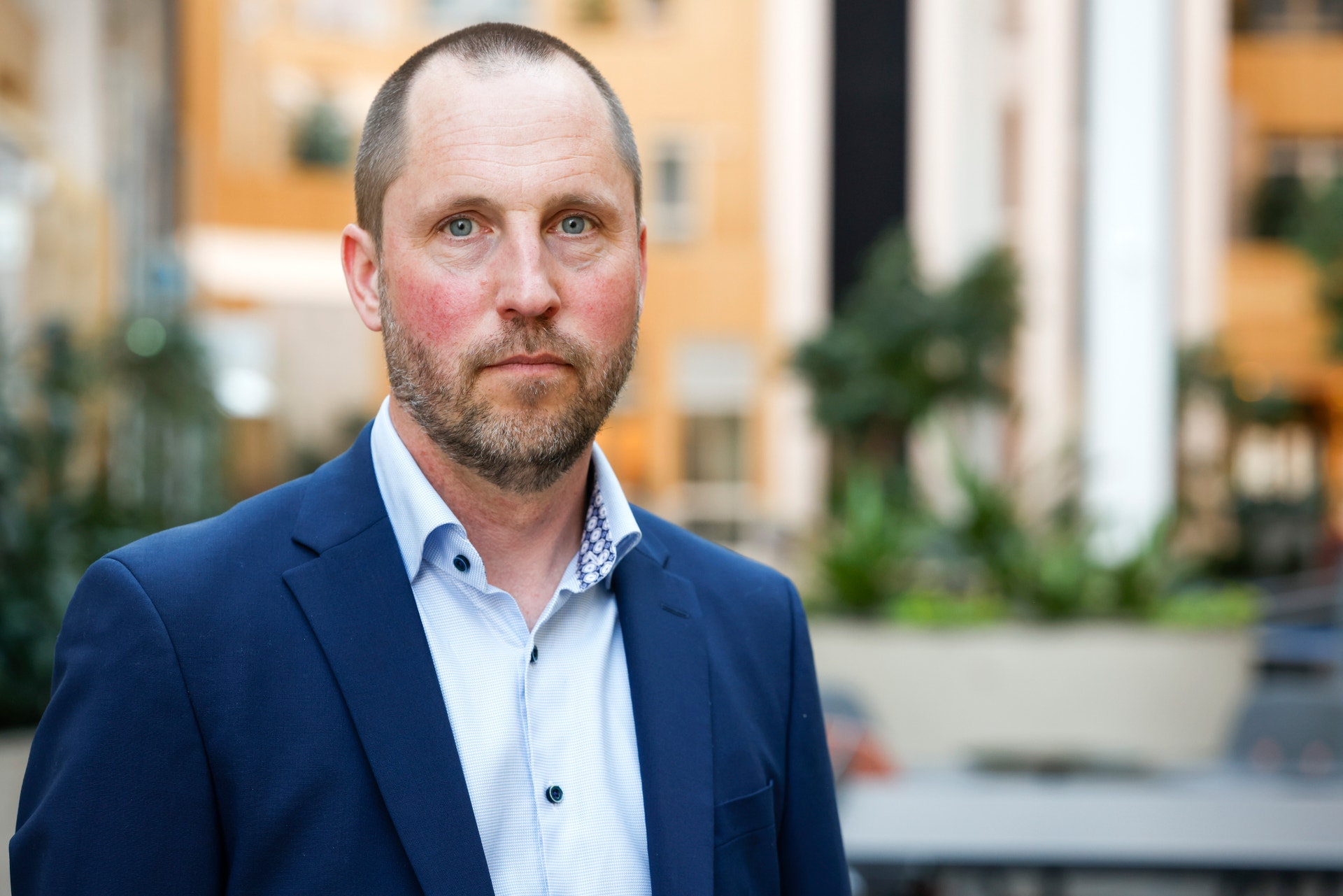 Porträtt av en man med kort hår och skägg, klädd i mörkblå kavaj och ljus skjorta, som står utomhus och tittar allvarligt in i kameran.