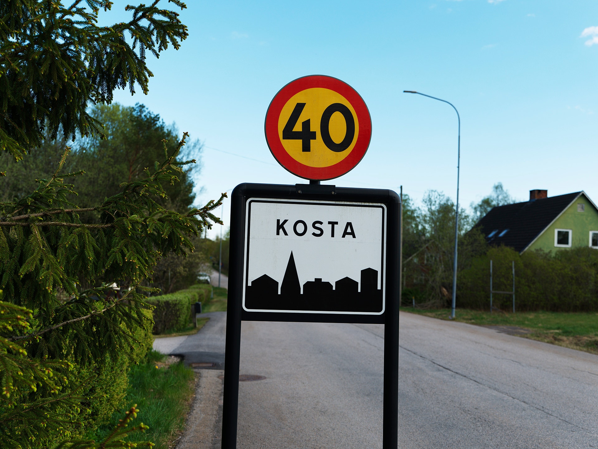 Vägskylt för Kosta med hastighetsbegränsning 40 km/h vid en väg omgiven av gröna träd och hus.