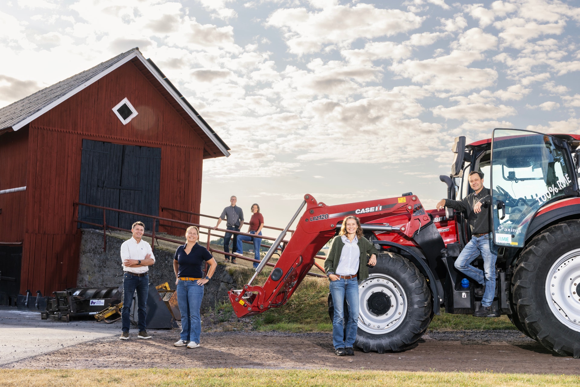 Fem personer står utomhus vid en röd ladugård och en röd traktor en solig dag.