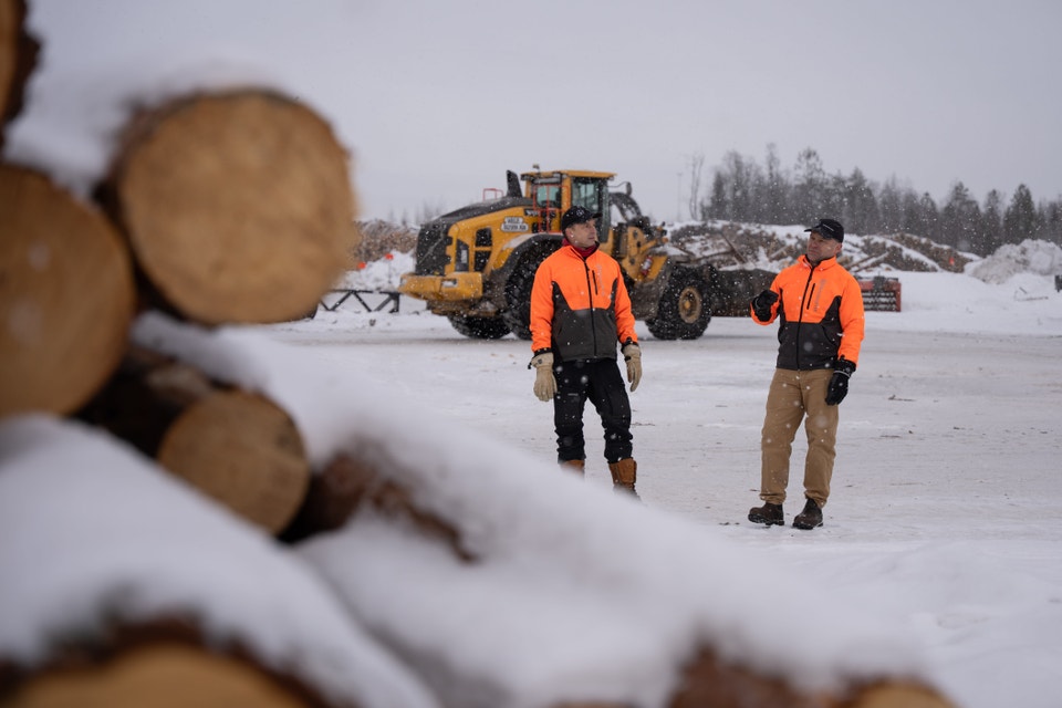 Två arbetare i orange reflektvester står på snöig mark vid en gul lastmaskin på en industriell terminal.