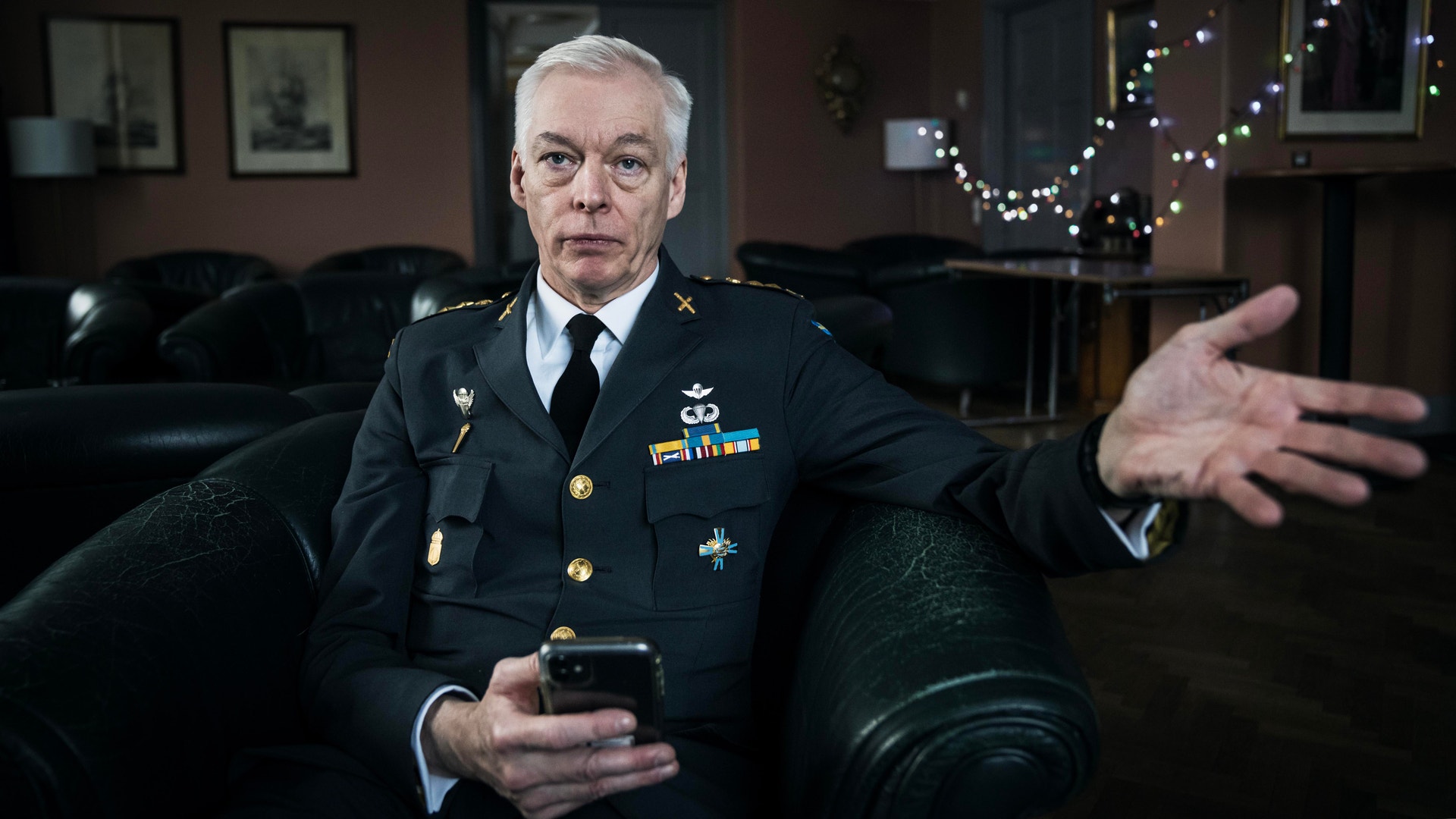 En äldre man i militär uniform sitter i en mörkare miljö. Han har grått hår och bär en mörk uniform med medaljer. Bakom honom syns juldekorationer.