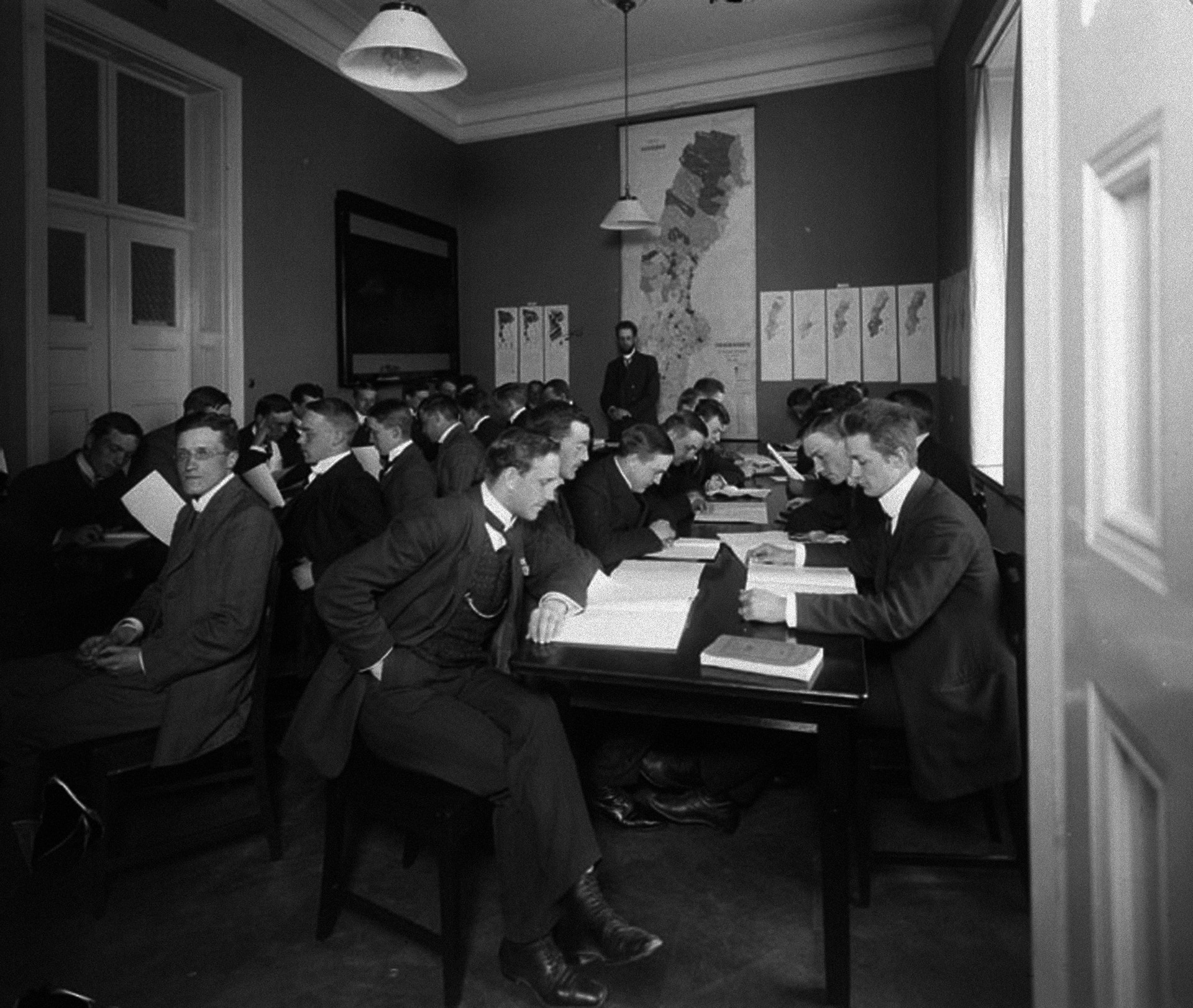 Svartvit historisk fotografi från cirka 1900-talet som visar ett klassrum med många män i mörka kostymer sittande vid bord med dokument. En instruktör står vid en tavla i bakgrunden.