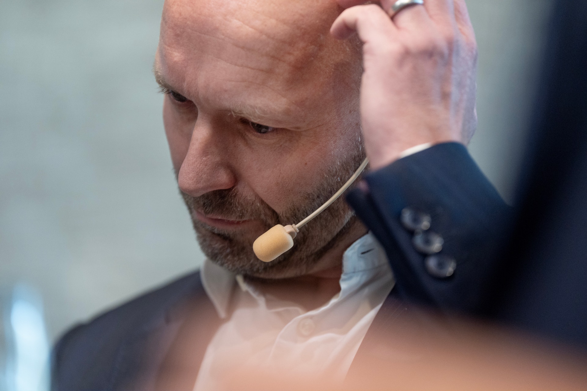 Närbild på en kal man med skägg och headset som blickar nedåt med ena handen mot huvudet. Han bär mörk kavaj och vit skjorta.
