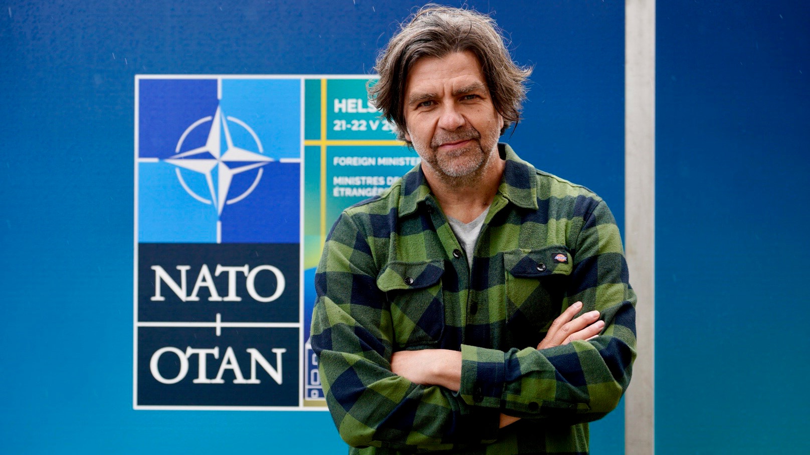Man i grön ruttig skjorta står framför NATO-logotyp på blå bakgrund.