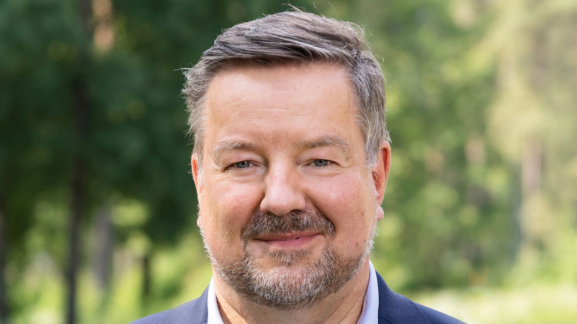 Porträtt av en man i mörkblå kavaj och ljusblå skjorta framför en grön naturlig bakgrund.