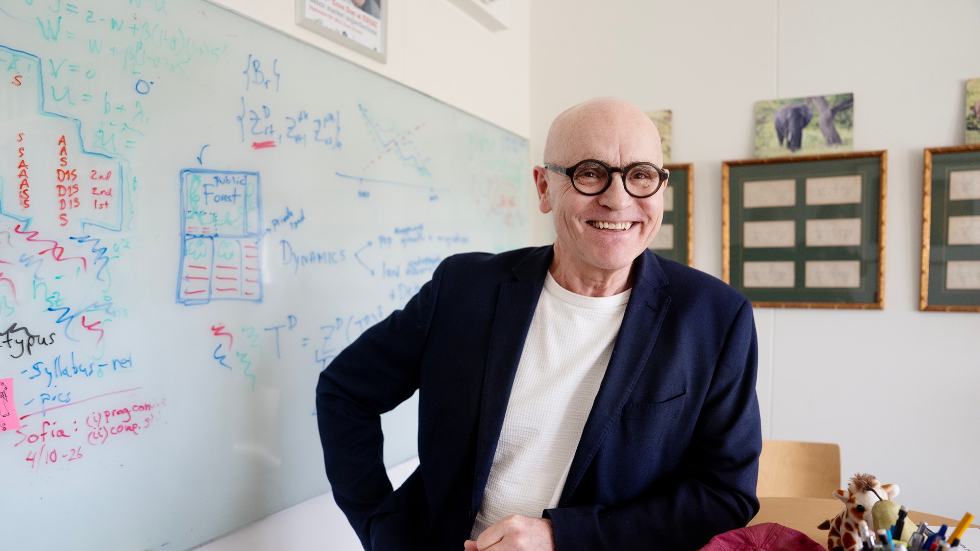 Man i glasögon och mörk kavaj poserar i ett arbetsrum med whiteboard i bakgrunden.