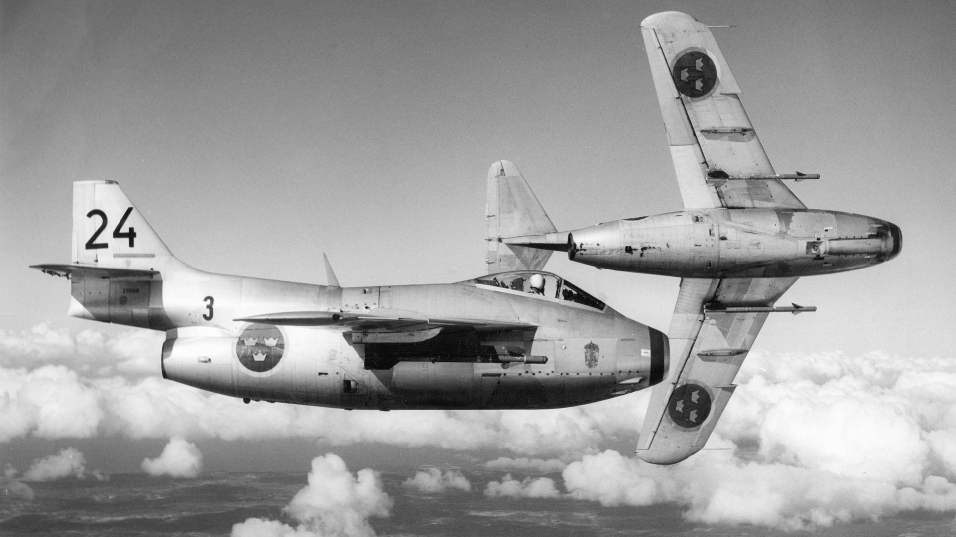 Svartvit flygfoto av ett klassiskt militärflygplan från 1940-50-talet som flyger över molnen. Planet har nummer 24 och 3 på sidorna.