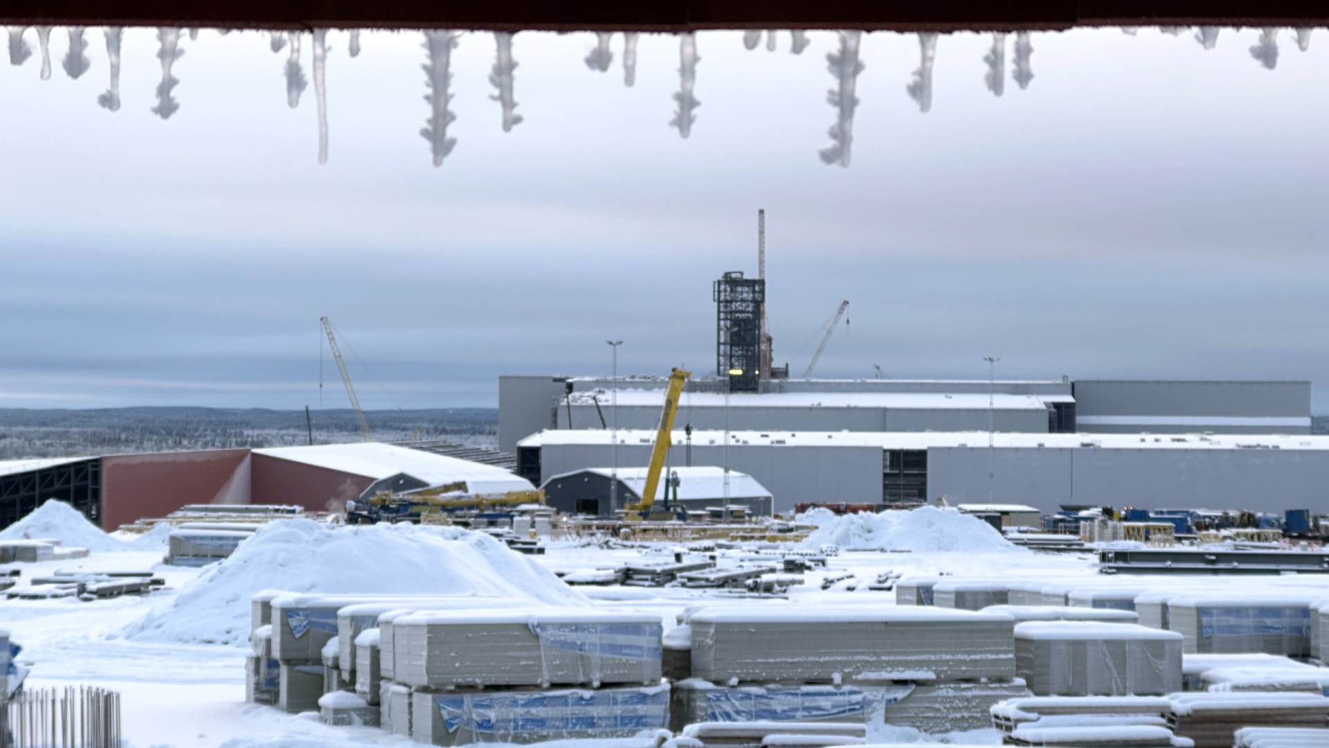 Snötäckt industriområde med byggmaterial, kranar och fabriksbyggnader i vintermiljö