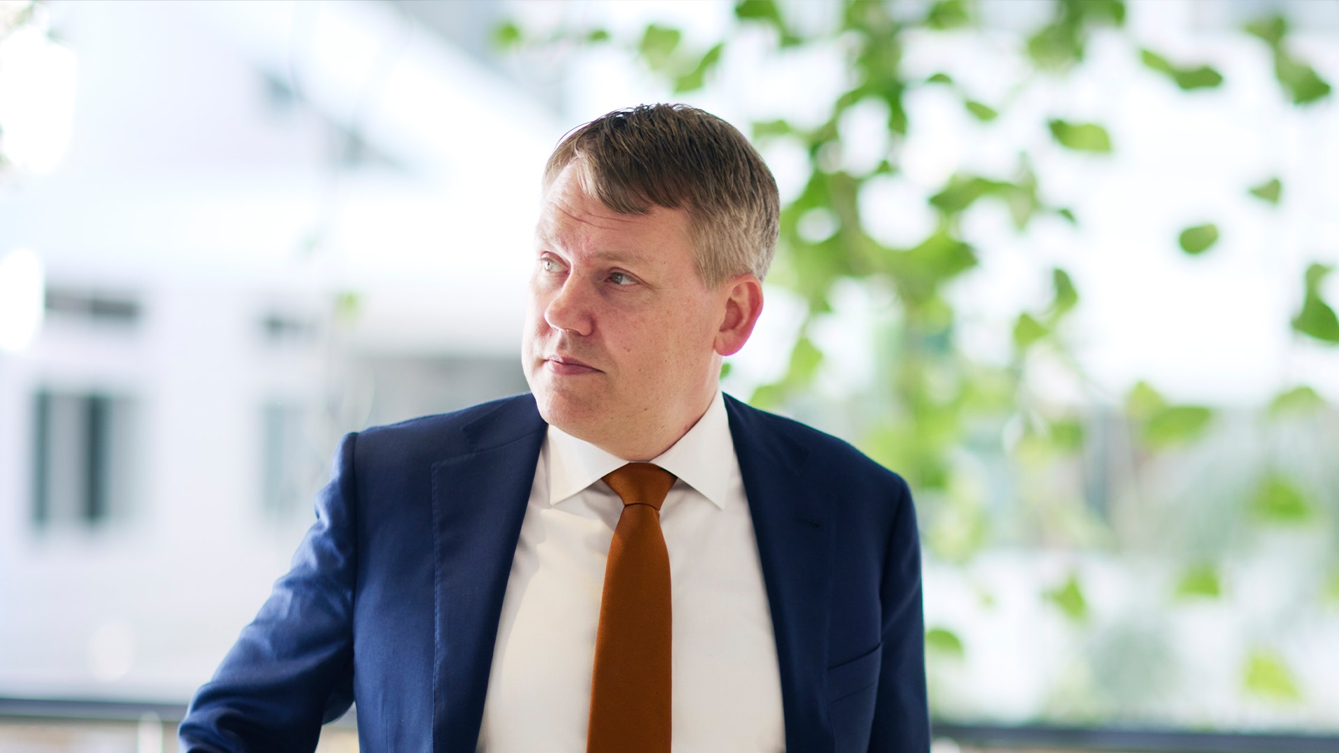 Man i mörkblå kostym och brun slips framför ett fönster med grön växtlighet i bakgrunden.