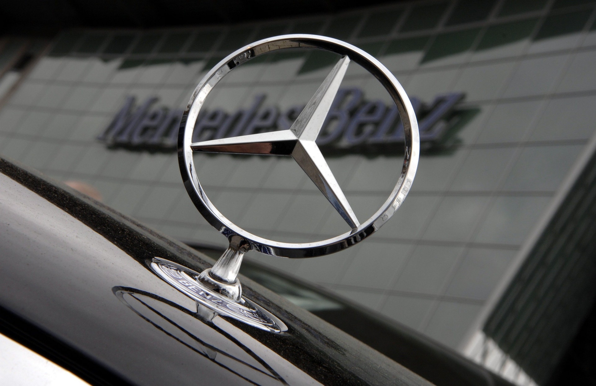 Mercedes-stjärnan på huven av en svart bil, framför en byggnad med Mercedes-Benz-skylt.
