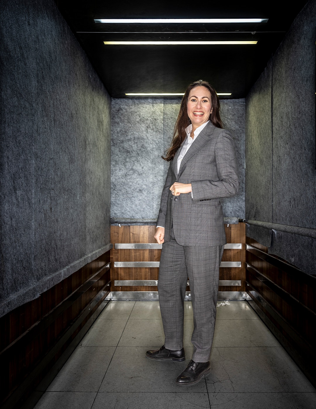 Professionell kvinna i grå kostym står i en modern hiss med mörkgrå väggar, träpaneler och LED-belysning. Hon ler mot kameran med en självsäker hållning.