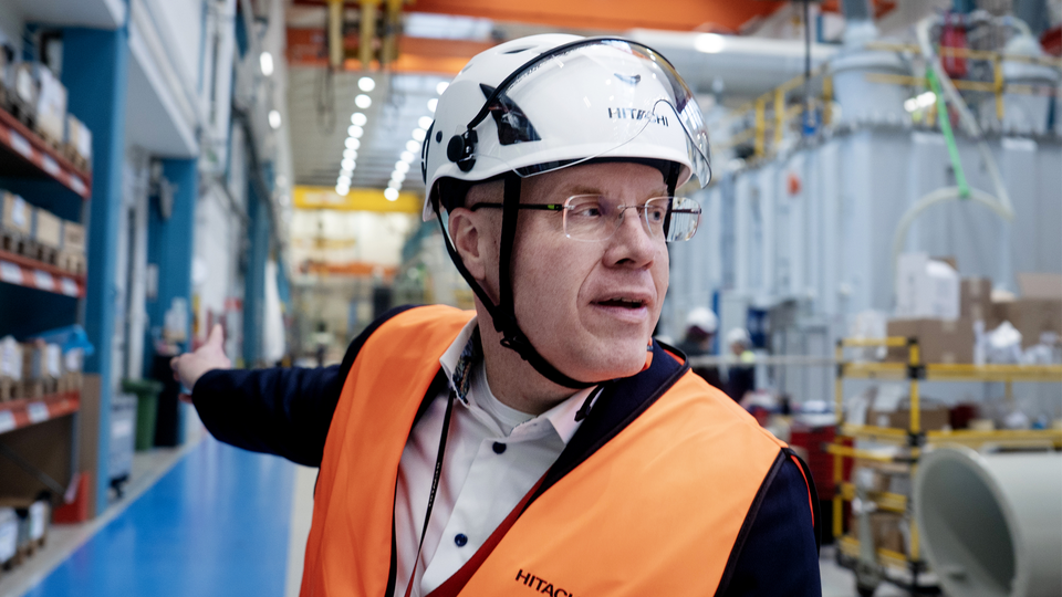 En man med glasögon och vit säkerhetshjalm står i en industriell fabriksmiljö. Han bär en orange och mörkblå arbetsvest med märkning och en vit skjorta. I bakgrunden syns industriell utrustning, lagerhyllor med material och gul kranutrustning. Mannen pekar eller gestikulerar medan han verkar kommunicera något.