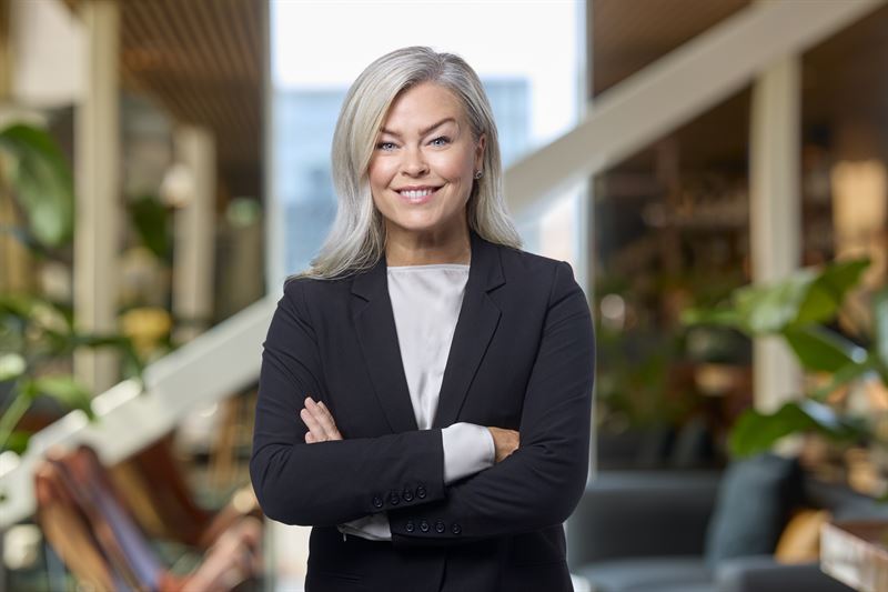 Porträtt av en professionell kvinna med gråvitt hår, iklädd svart blazer och vit skjorta, som står med armarna i kors och ler mot kameran i ett modernt kontorslandskap.