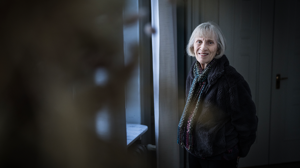 Porträttfoto av Claudia Goldin med vitt hår, iklädd en svart jacka och mönstrad scarf, som står i ett modernt inredningsrum och ler mot kameran.