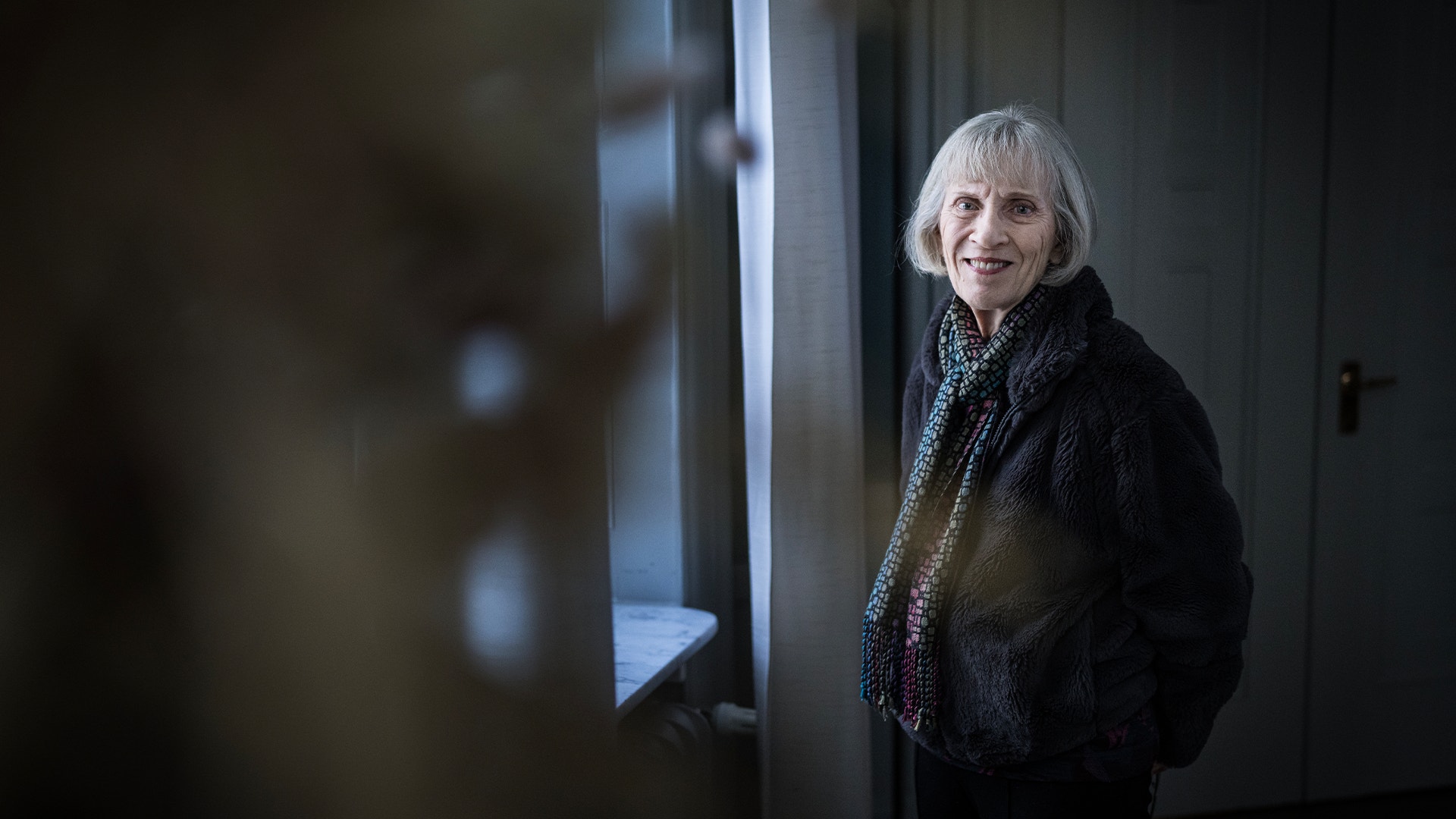 Porträttfoto av Claudia Goldin  med vitt hår, iklädd en svart jacka och mönstrad scarf, som står i ett modernt inredningsrum och ler mot kameran.