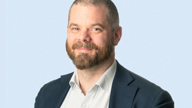 Porträtt av en man med kort hår och skägg, klädd i mörkblå kavaj och vit skjorta mot ljusblå bakgrund.